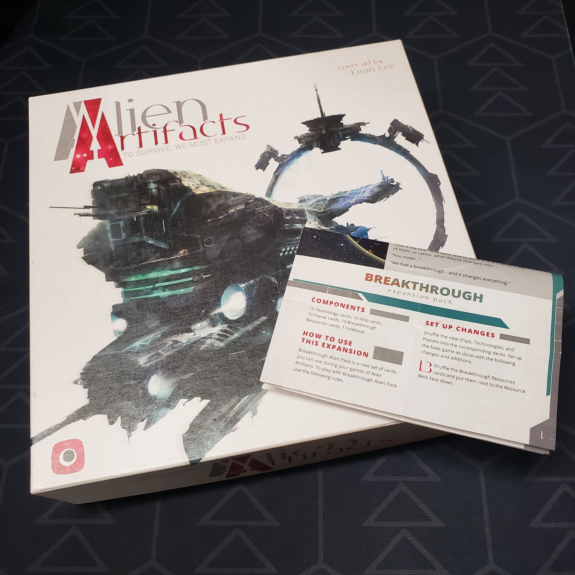 Image shows the front cover of the box of the Alien Artifacts board game with the instructions for the Breakthrough expansion sitting on top of it