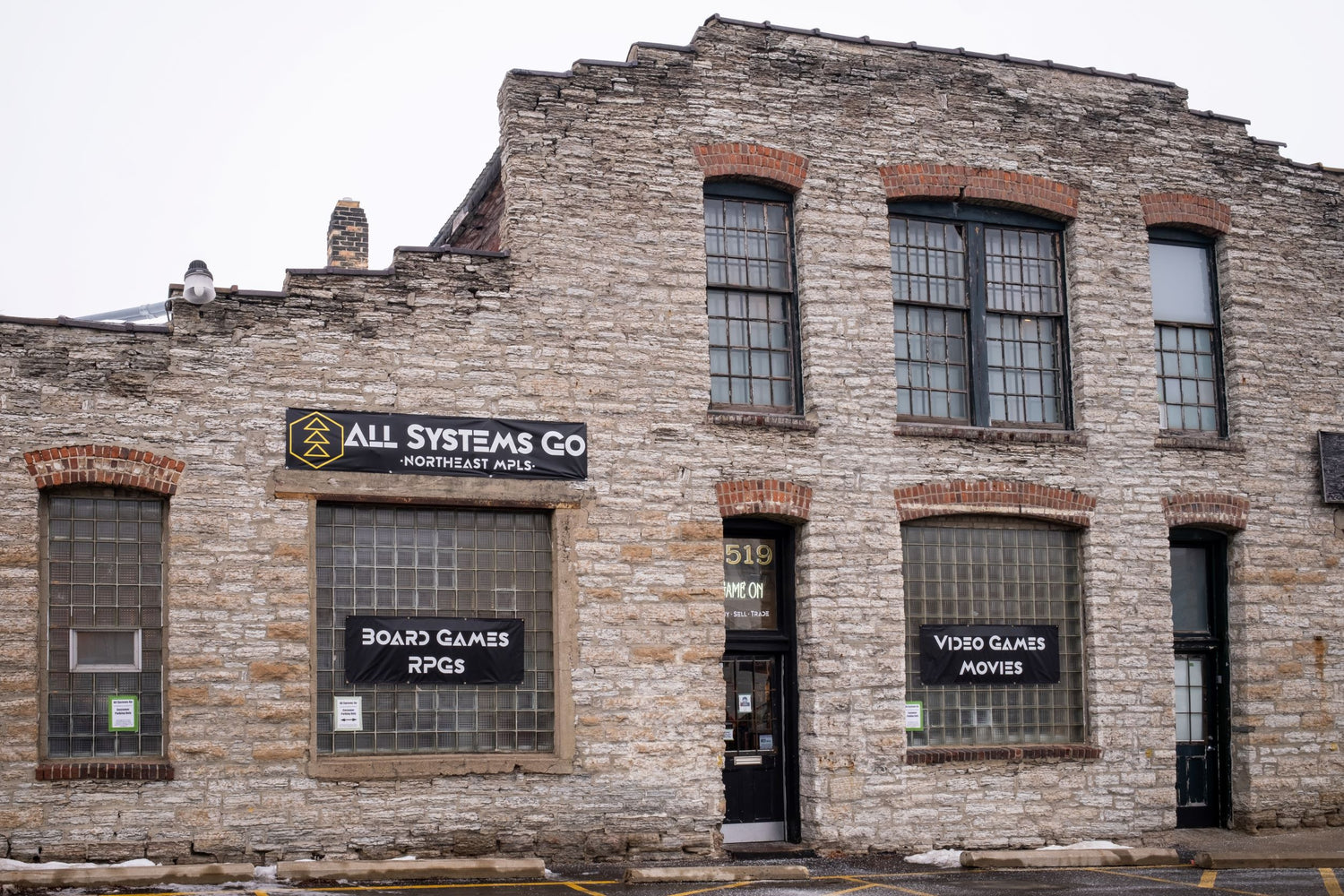 Image shows the front of the All Systems Go storefront in northeast Minneapolis. The building is brick and has 3 banners on it; reading the store name and the various items sold there.