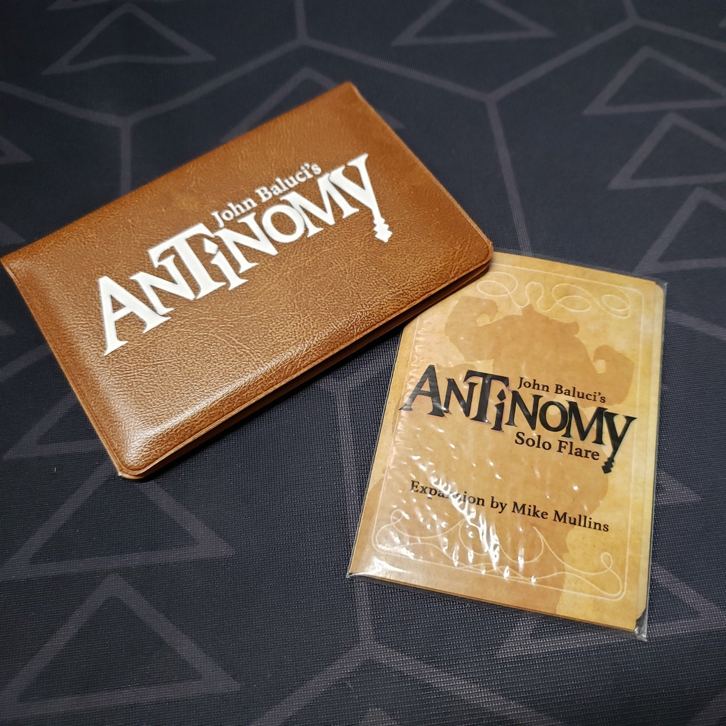 Image shows the front of the card wallet for the Antinomy card game wit the Solo Flare expansion sitting next to ti