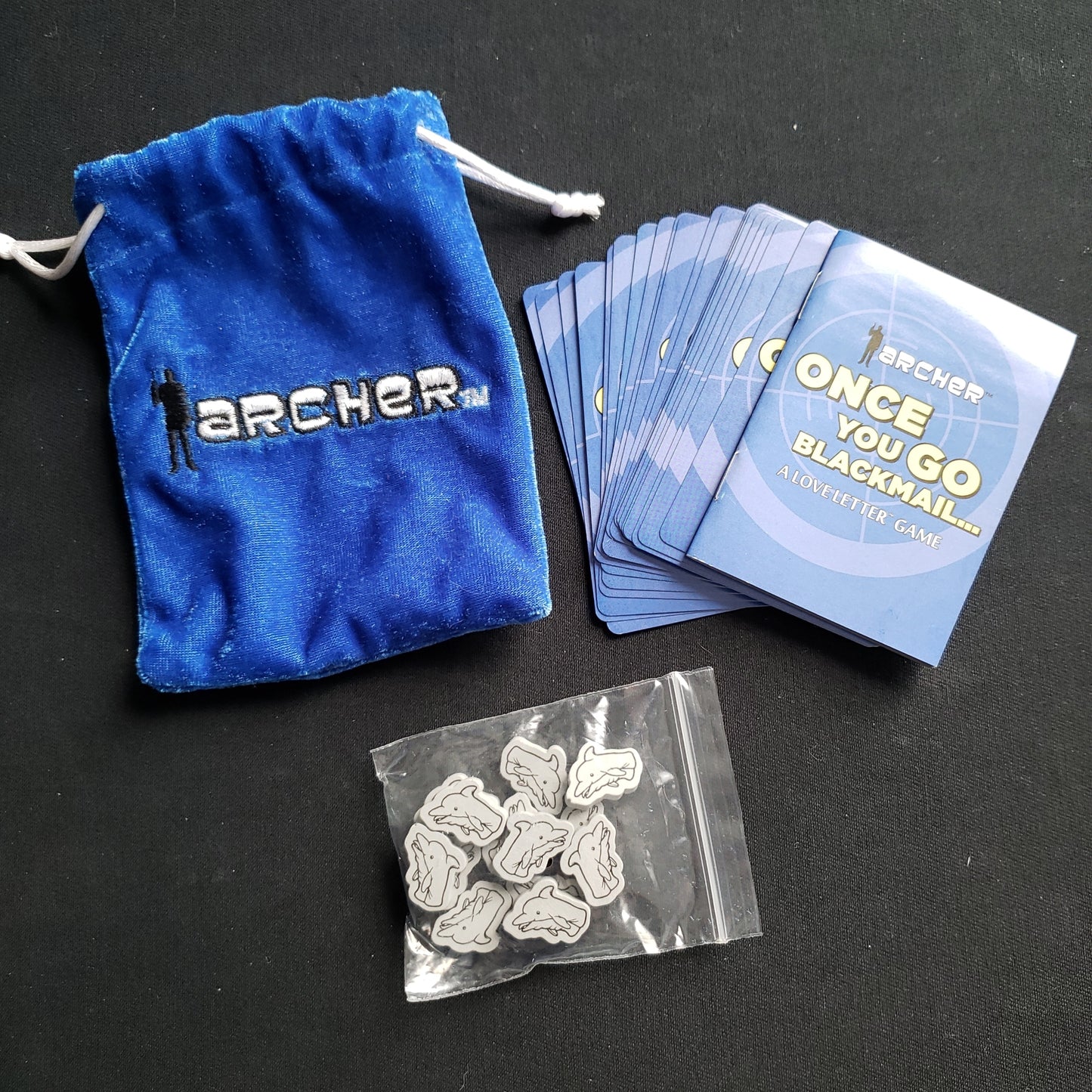 Image shows the components & carrying bag fanned out for the card game Archer: Once You Go Blackmail