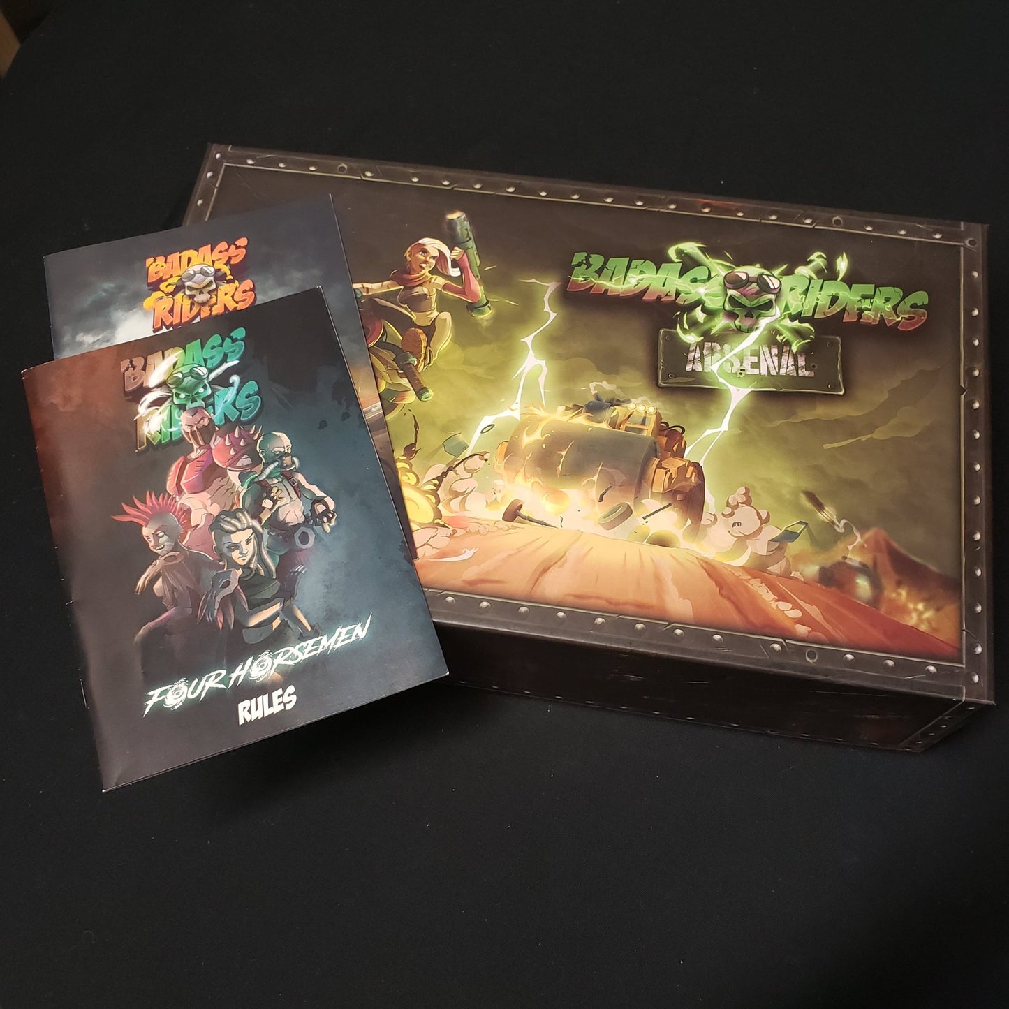 Image shows the front cover of the Arsenal box of the Badass Riders board game, with two instruction manuals sitting on top of it