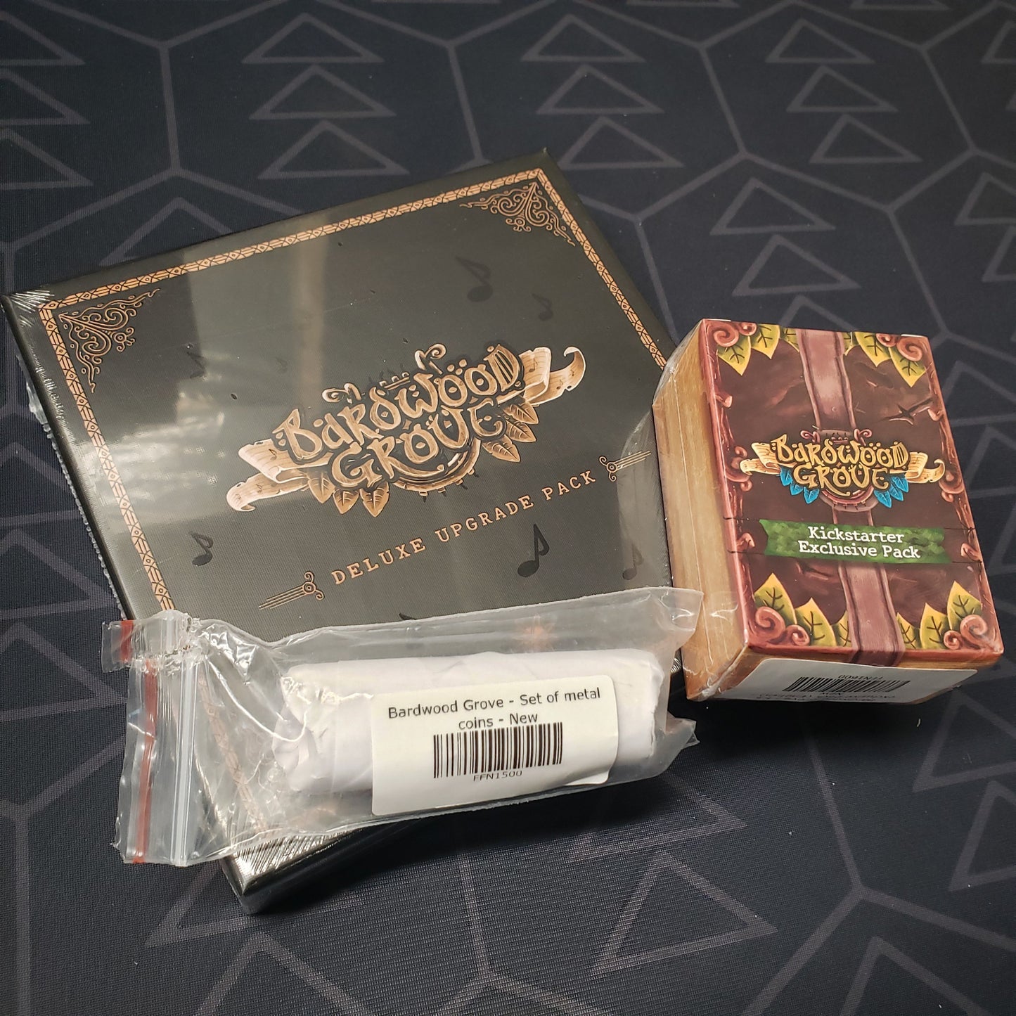 Image shows the front cover of the packages of the Deluxe Upgrade Pack, Kickstarter Exclusives + Metal Coins packs for the board game Bardwood Grove