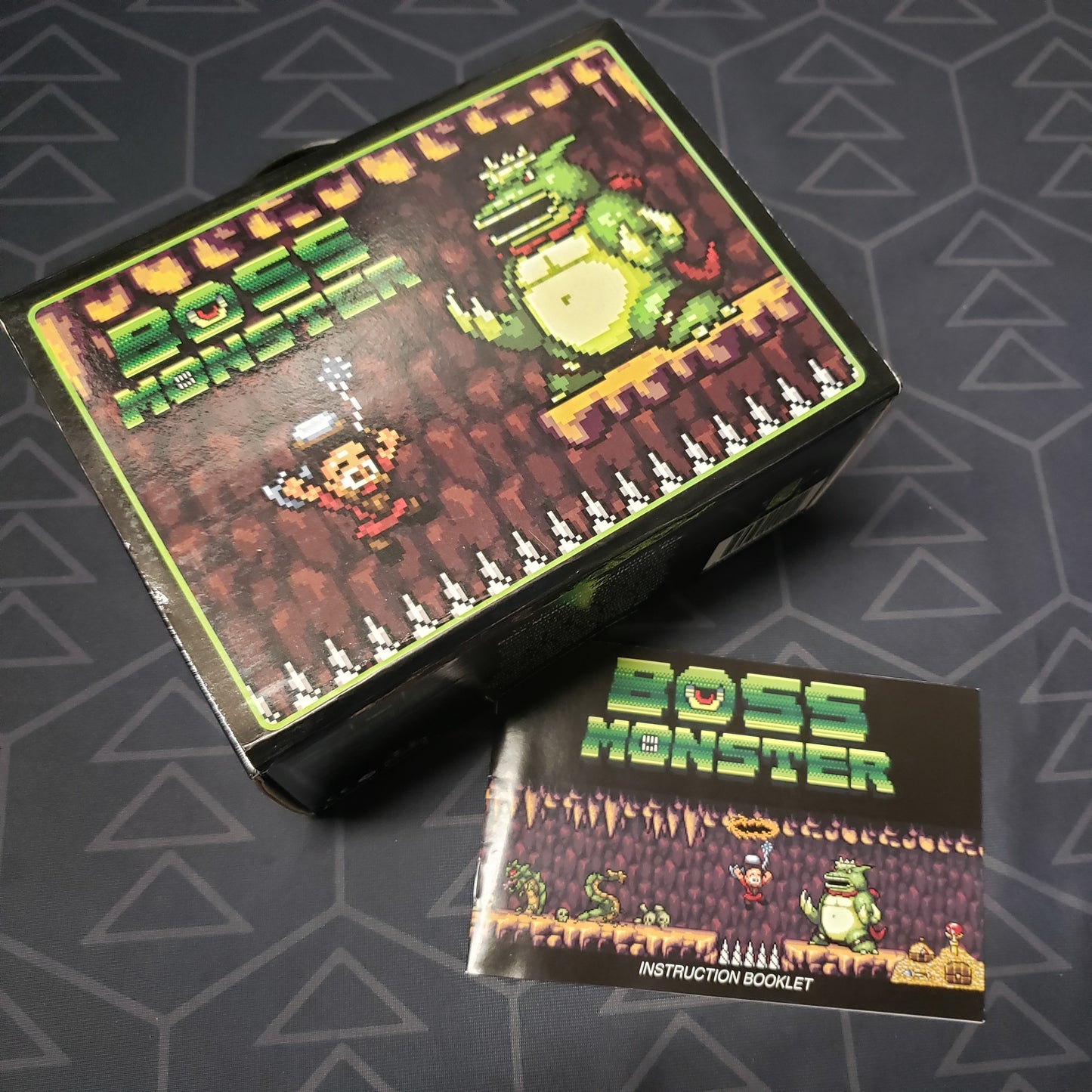 Image shows the front cover of the Collector Box for the card game Boss Monster, sitting next to an instruction manual
