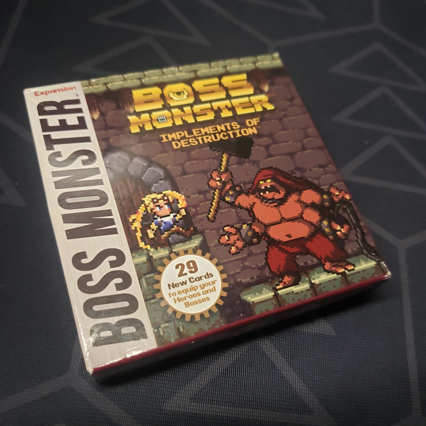 Image shows the front cover of the box of the Implements of Destruction expansion for the card game Boss Monster
