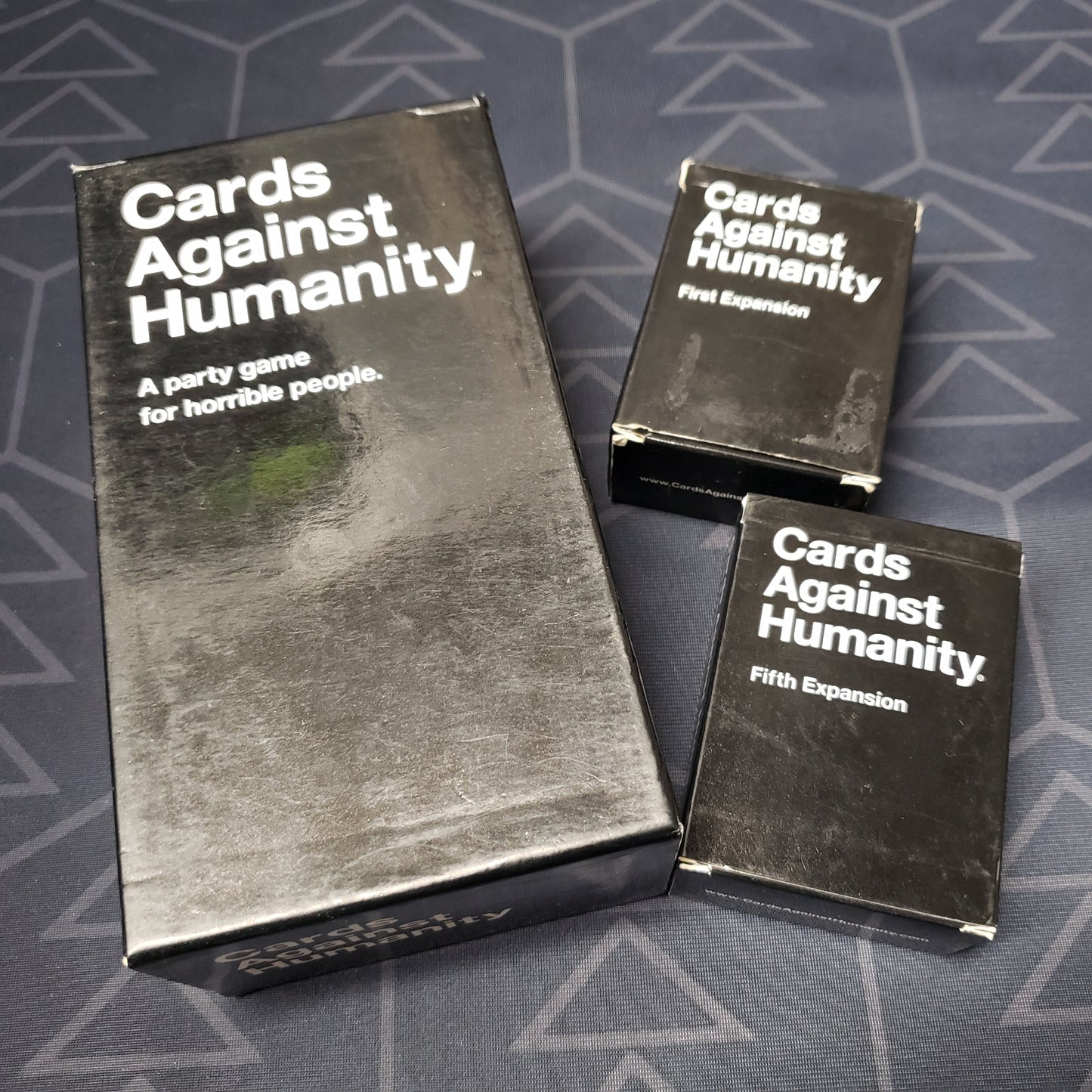Image shows the front cover of the box for the Cards Against Humanity card game, that is sitting next to the boxes of the First and Fifth expansions