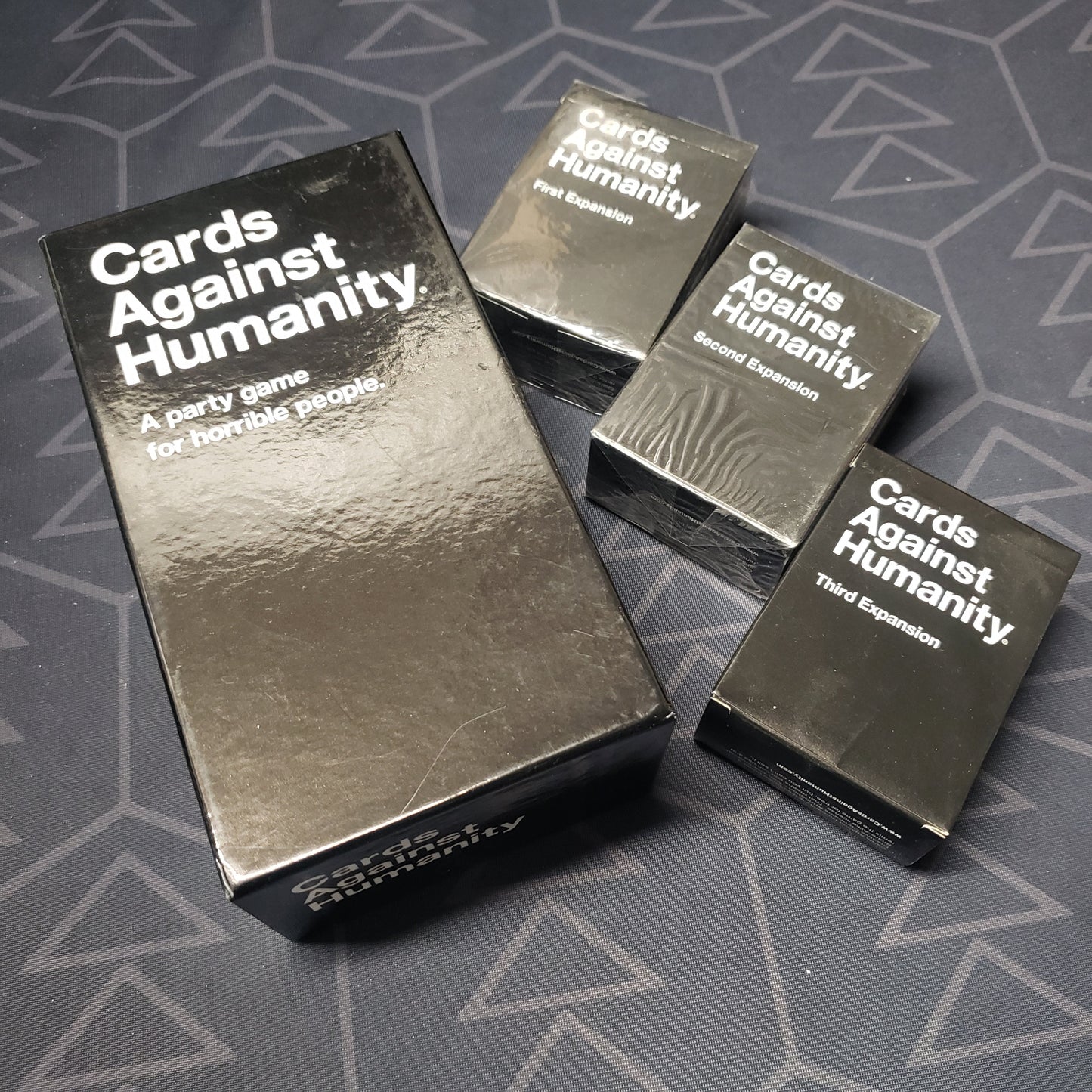 Image shows the front cover of the box for the Cards Against Humanity card game, that is sitting next to the boxes of the First, Second and Third expansions