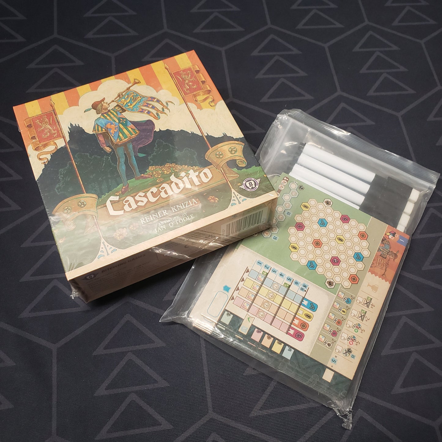 Image shows the front cover of the box of the Cascadito board game sitting on top of the dry erase boards for it