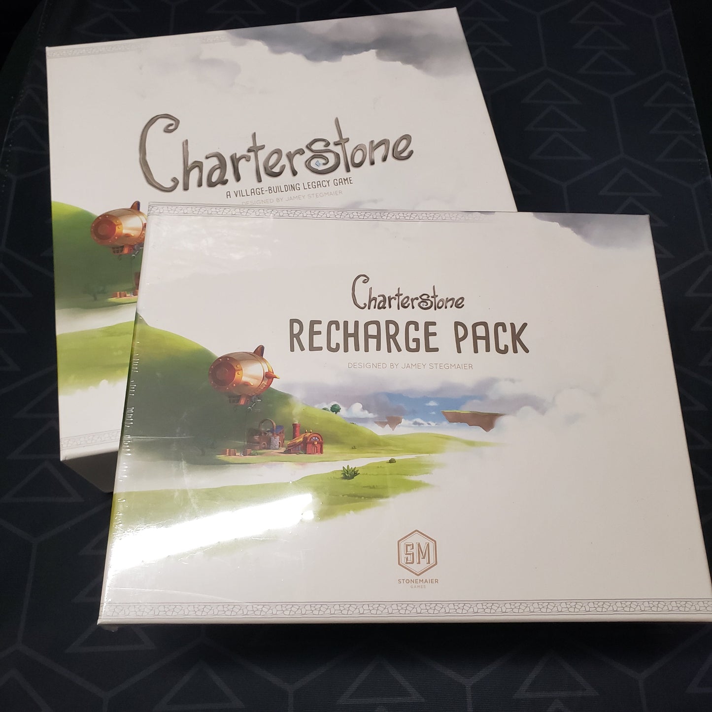 Image shows the front cover of the box for the Charterstone board game with the Recharge Pack for the game sitting on top of it