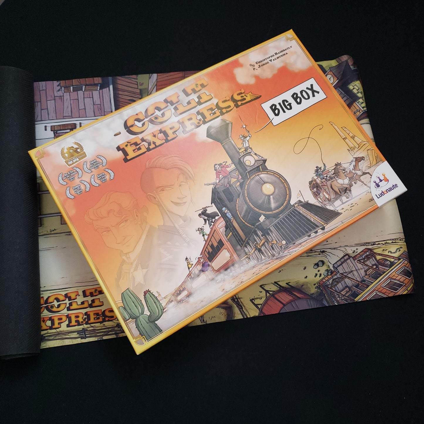 Image shows the front cover of the box of the Colt Express: Big Box board game, sitting on top of the roll-out neoprene playmat for the game
