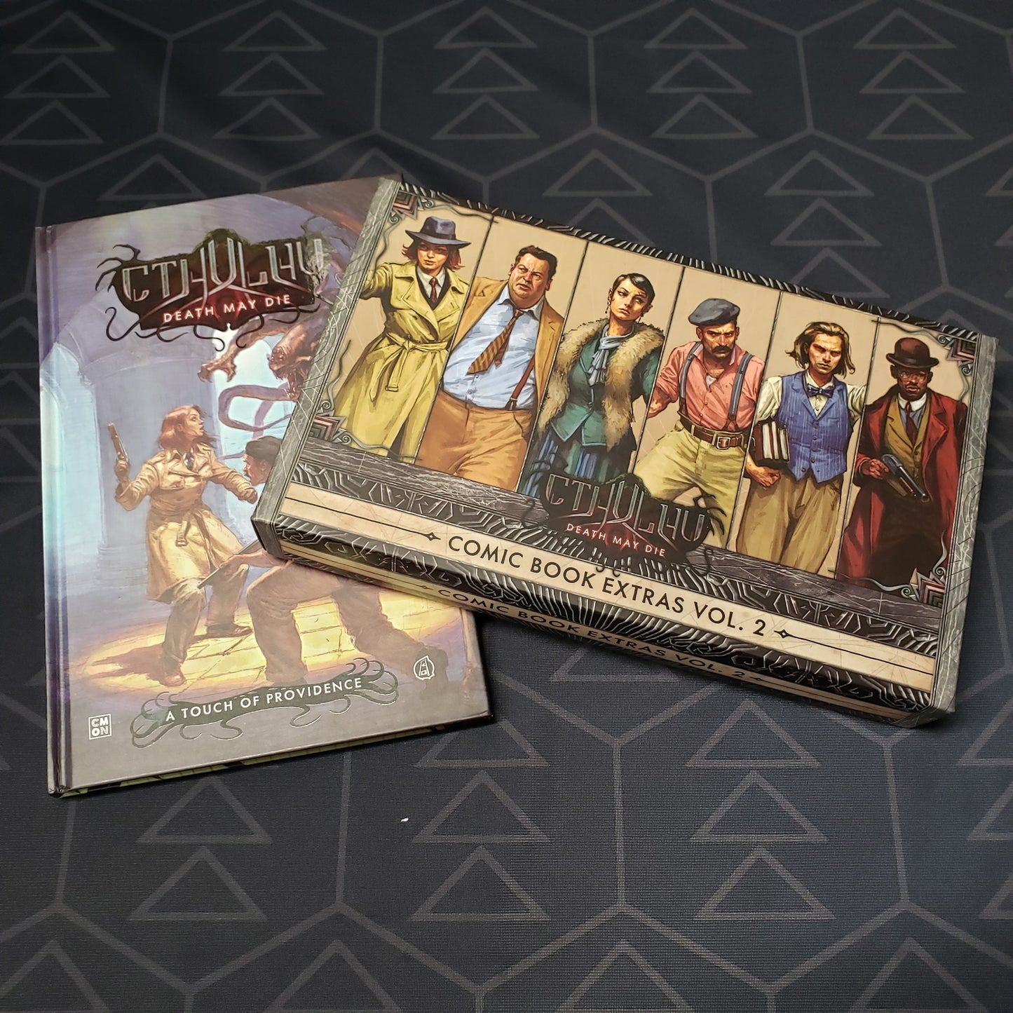 Image shows the front covers of the box for the Comic Book Extras Vol. 2expansion for the board game Cthulhu: Death May Die, as well as the "A Touch of Providence" graphic novel