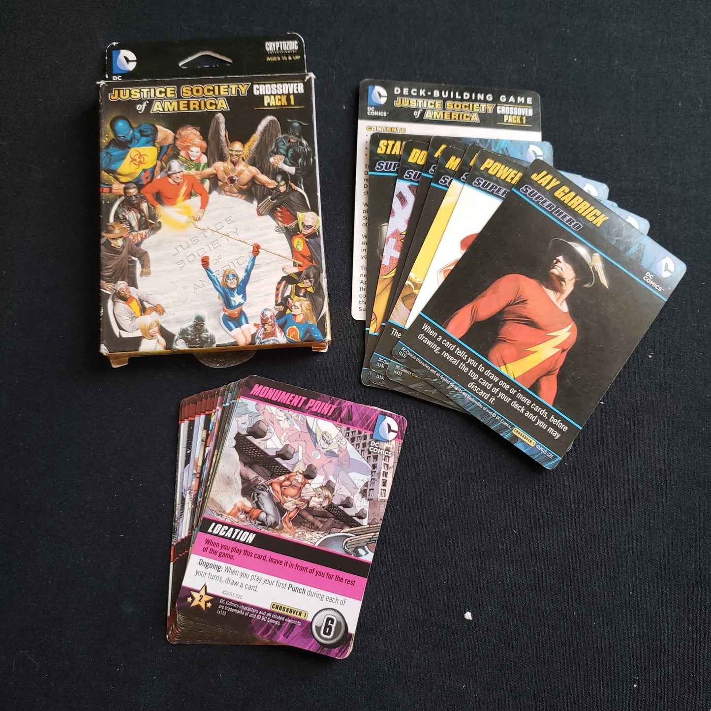 Image shows the front cover of the box of the Justice Society of America Crossover Pack expansion for the DC Comics Deckbuilding card game, along with the cards fanned out next to it