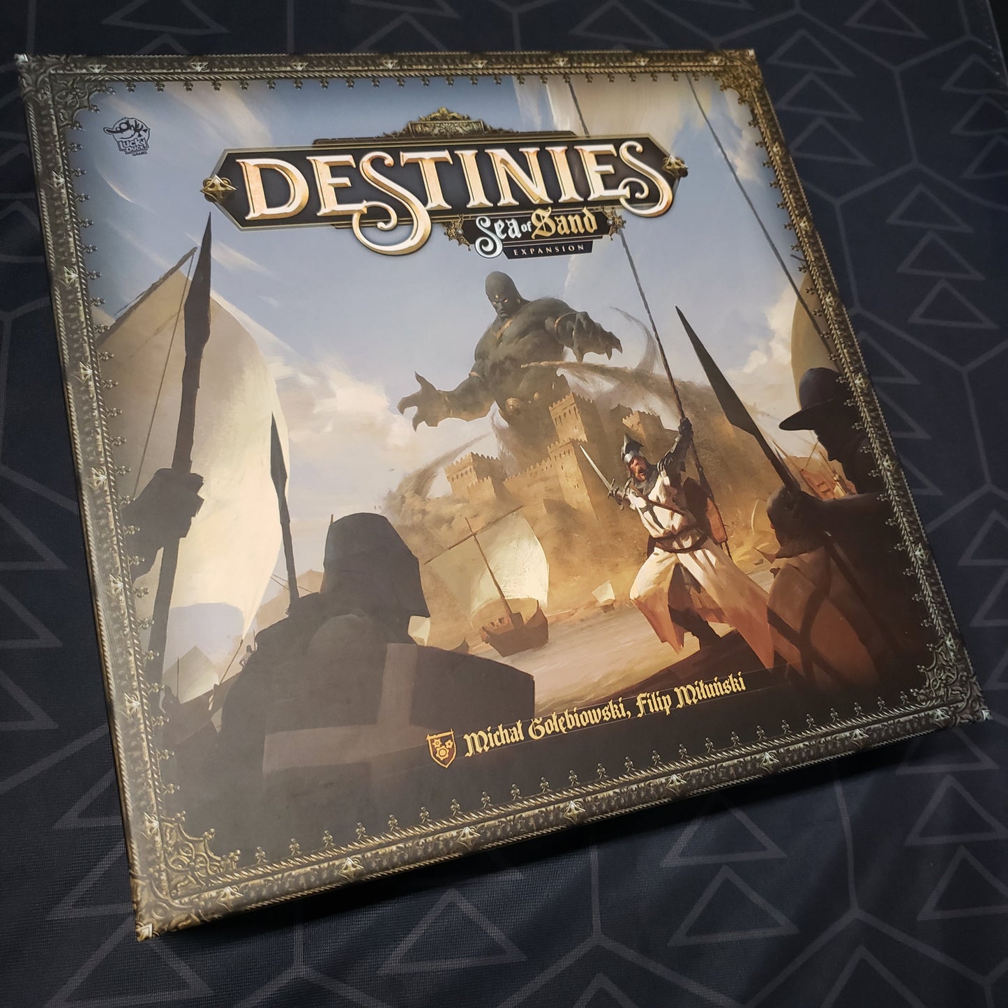 Image shows the front cover of the box of the Sea of Sand expansion for the board game Destinies