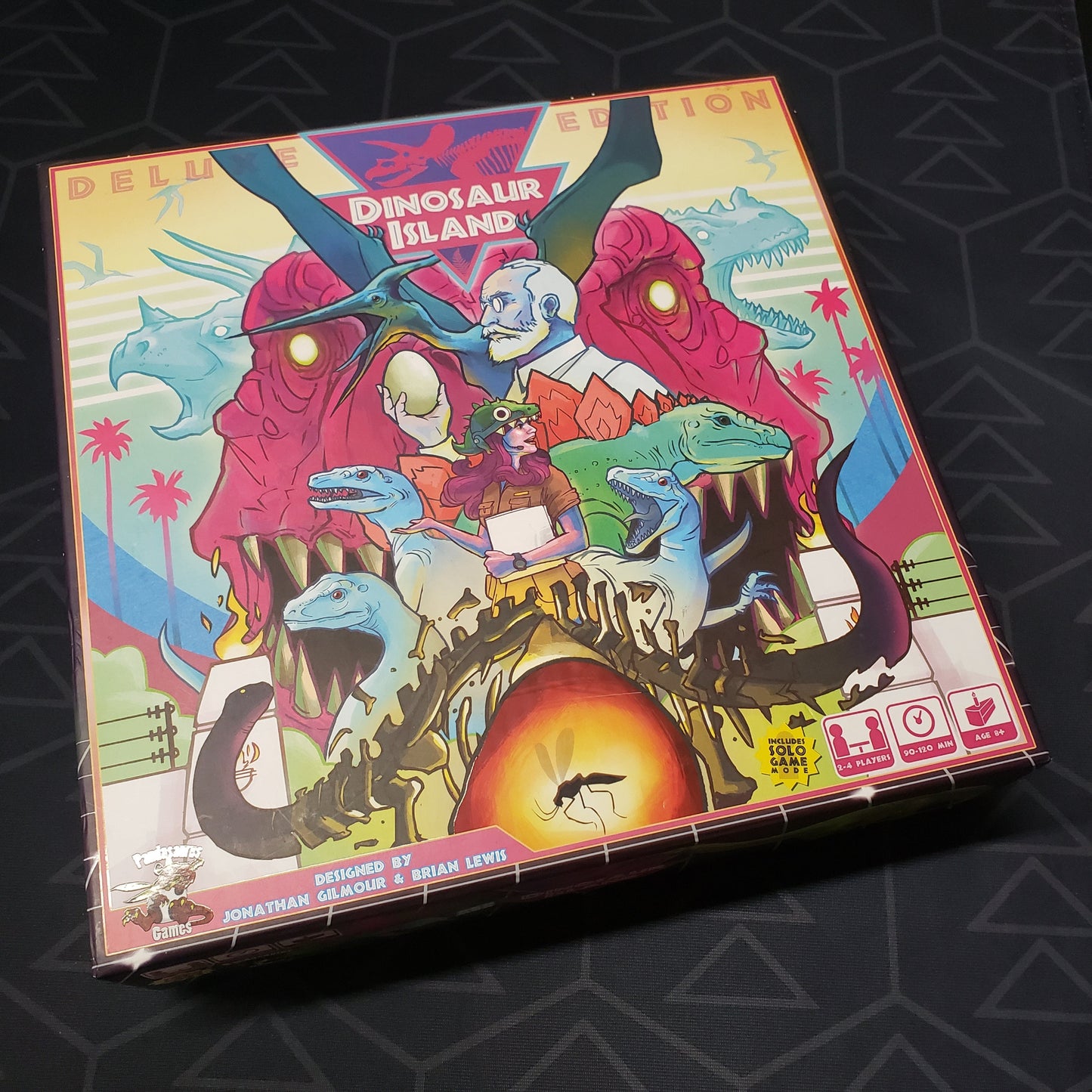 Image shows the front cover of the Deluxe Edition box of the board game Dinosaur Island