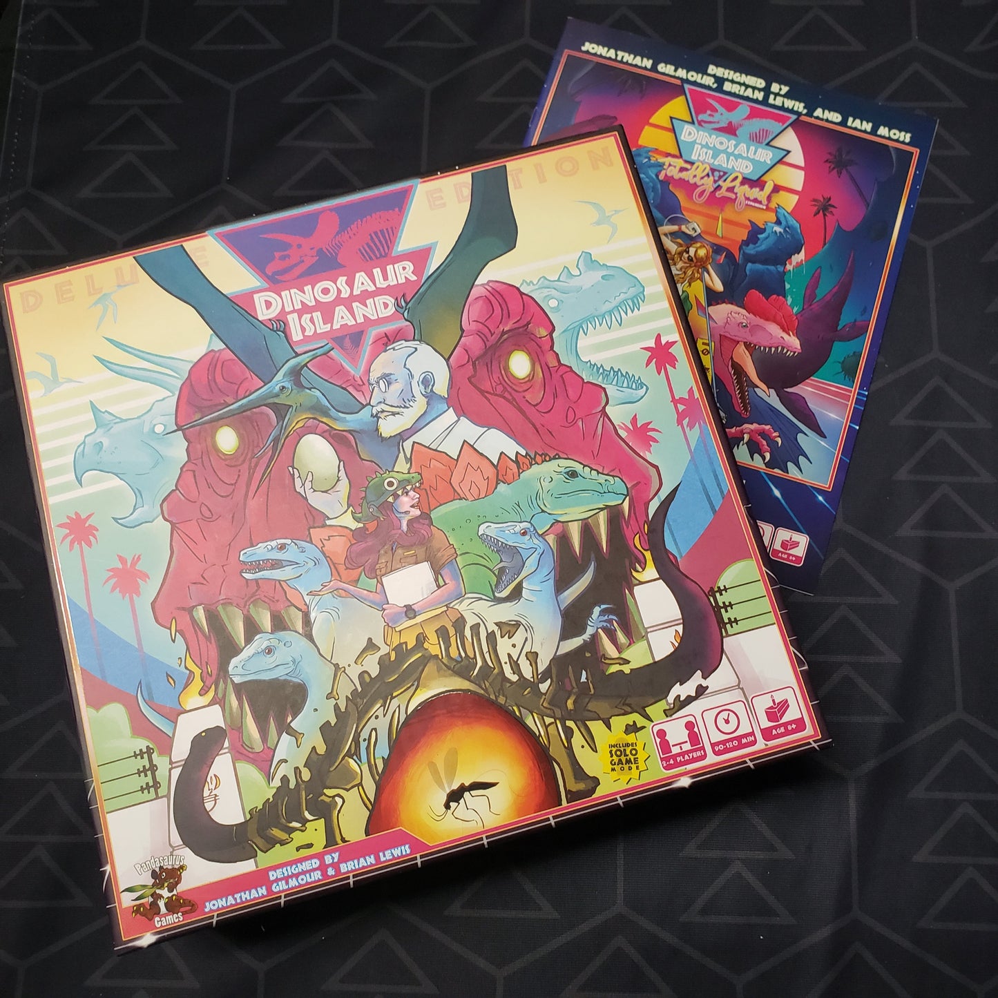 Image shows the front cover of the Deluxe Edition box of the board game Dinosaur Island sitting on top of the instructions for the Totally Liquid expansion