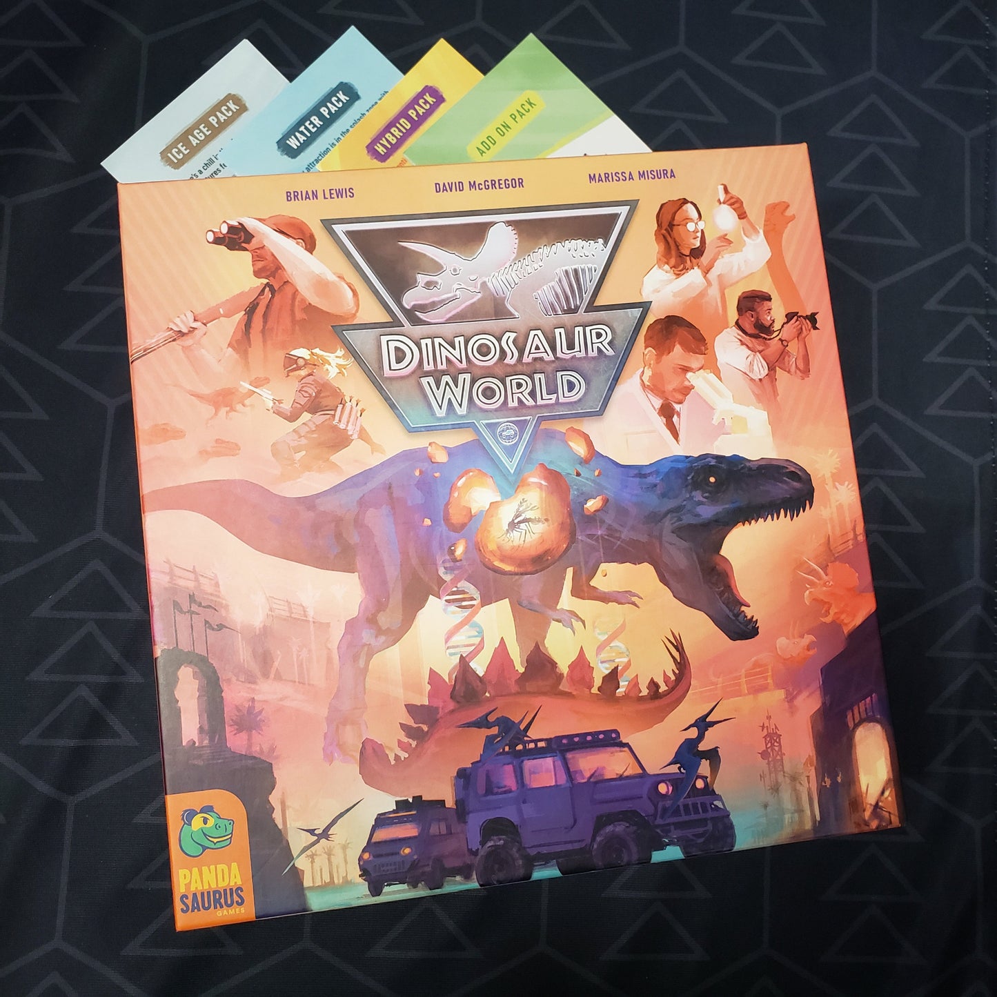 Image shows the front cover of the box for the Kickstarter Edition of the board game Dinosaur World sitting on top of the four instruction sheets for the included expansions