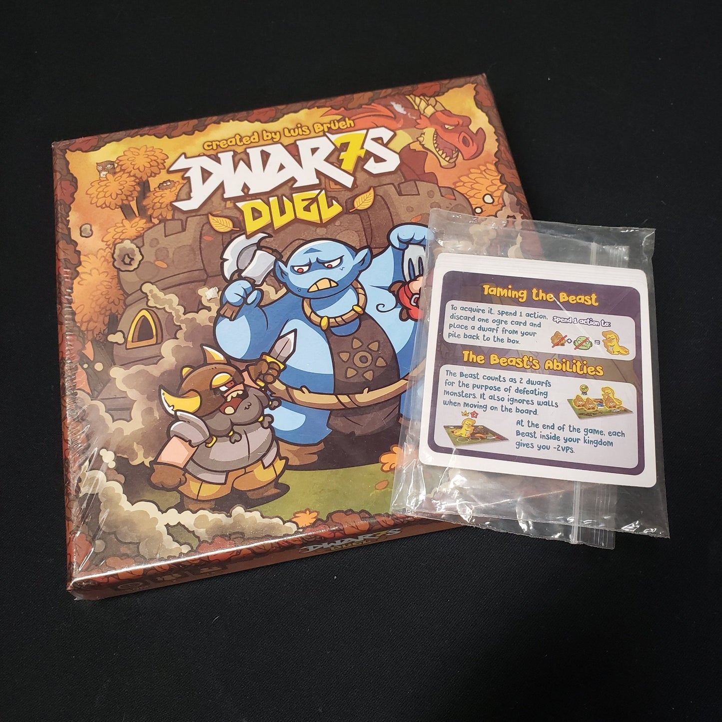 Image shows the front cover of the box of the Dwar7s Duel board game with a bagged promo pack of cards on top of it