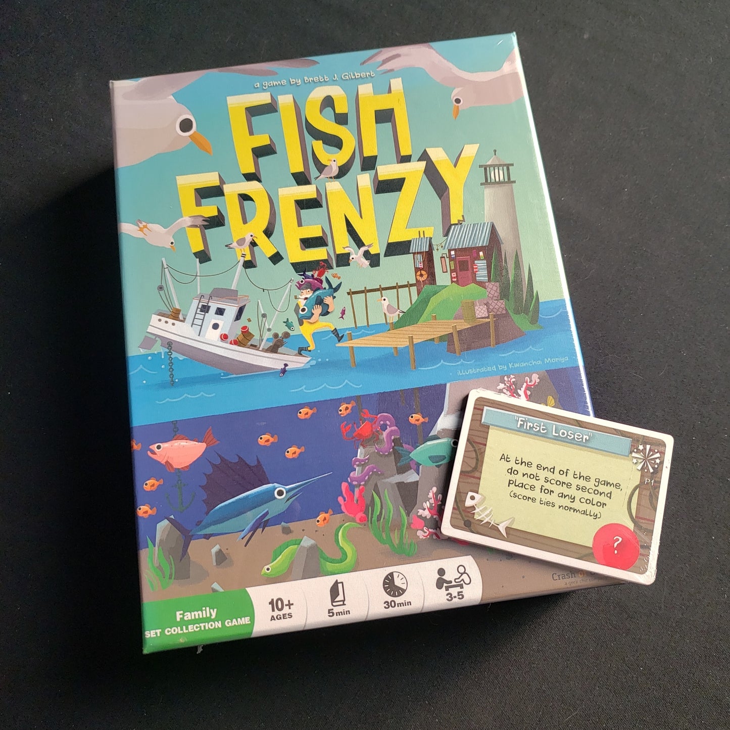 Image shows the front cover of the box of the Fish Frenzy board game with a promo card pack sitting on top of it