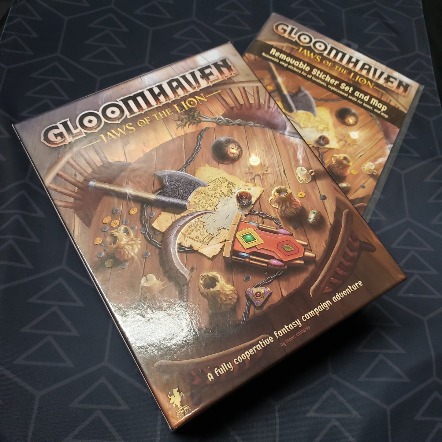 Image shows the front cover of the box of the Gloomhaven: Jaws of the Lion board game sitting on top of the Removeable Sticker Set & Map pack