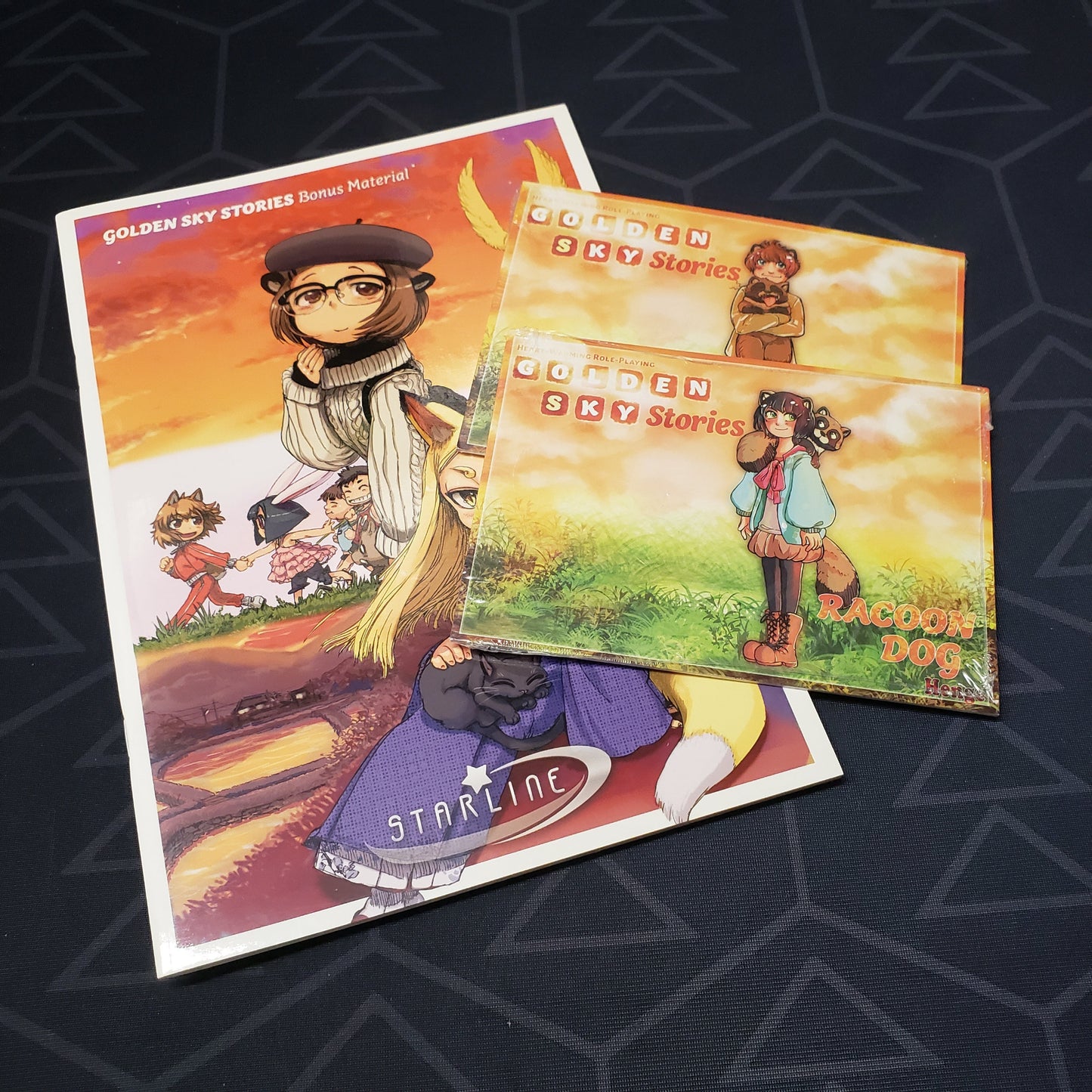 Image shows the front cover of the Colors of the Sky bonus book for the roleplaying game Golden Sky Stories, with two sealed packs of character cards sitting on top of it