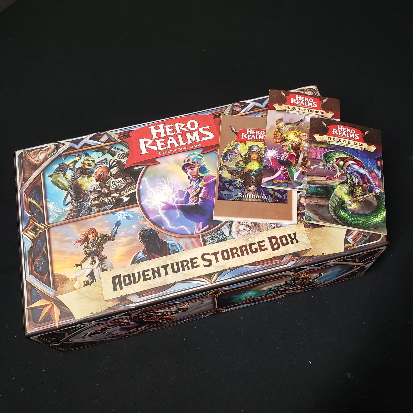 Image shows the front cover of the Adventure Storage Box for the card game Hero Realms, with 3 different rulebooks sitting on top of it