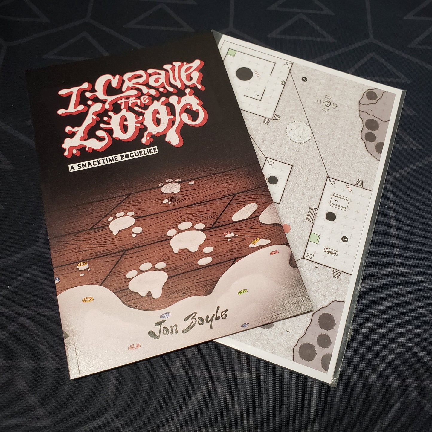 Image shows the front cover of the I Crave The Loop roleplaying game book, sitting on top of the map pack for the game