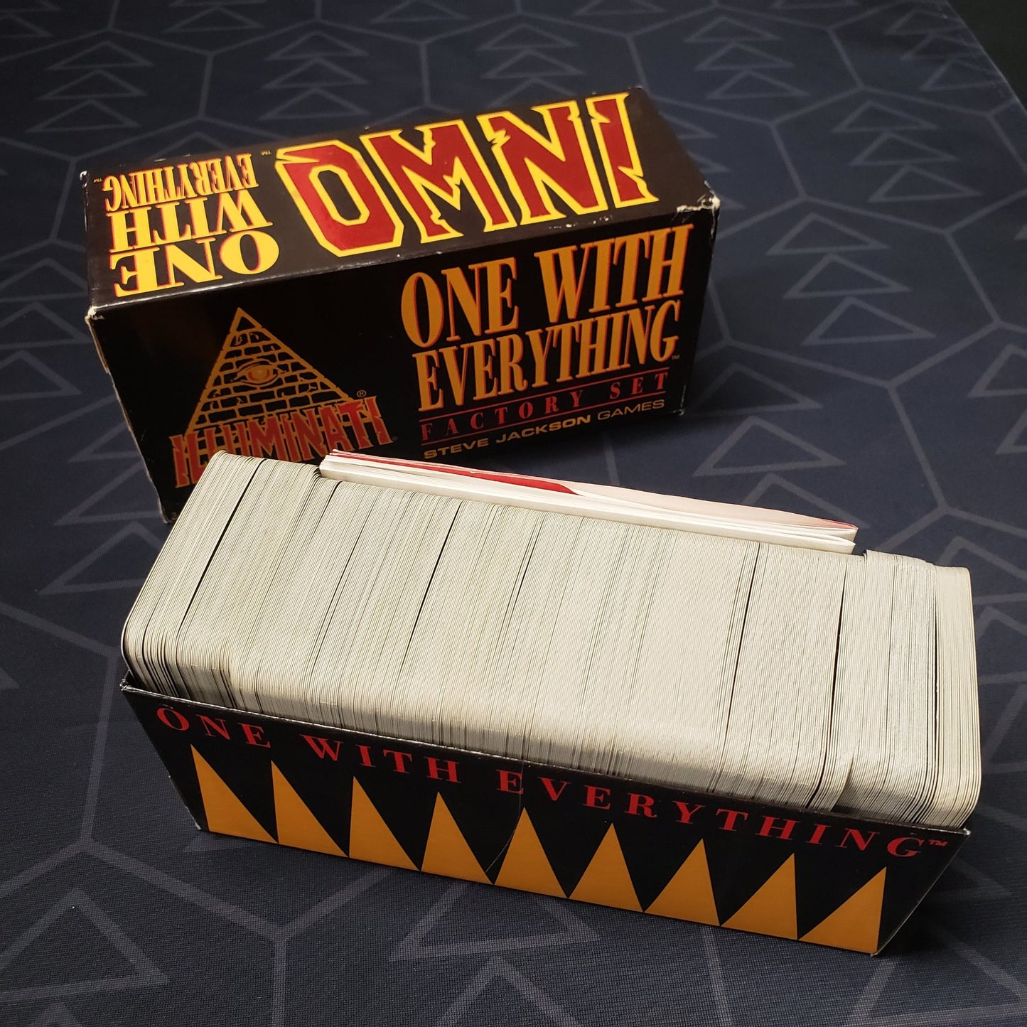 Image shows the top of the box of the One With Everything Factory Set for the card game Illuminati: new World Order, sitting next to the bottom of the box displaying the cards inside