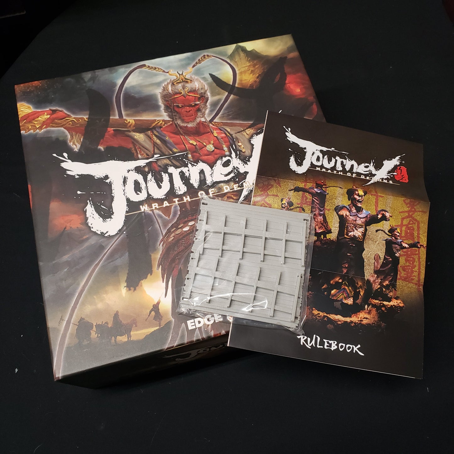 Image shows the front cover of the box of the Journey: Wrath of Demons board game, with expansion instructions and a bridge miniature sitting on top of it