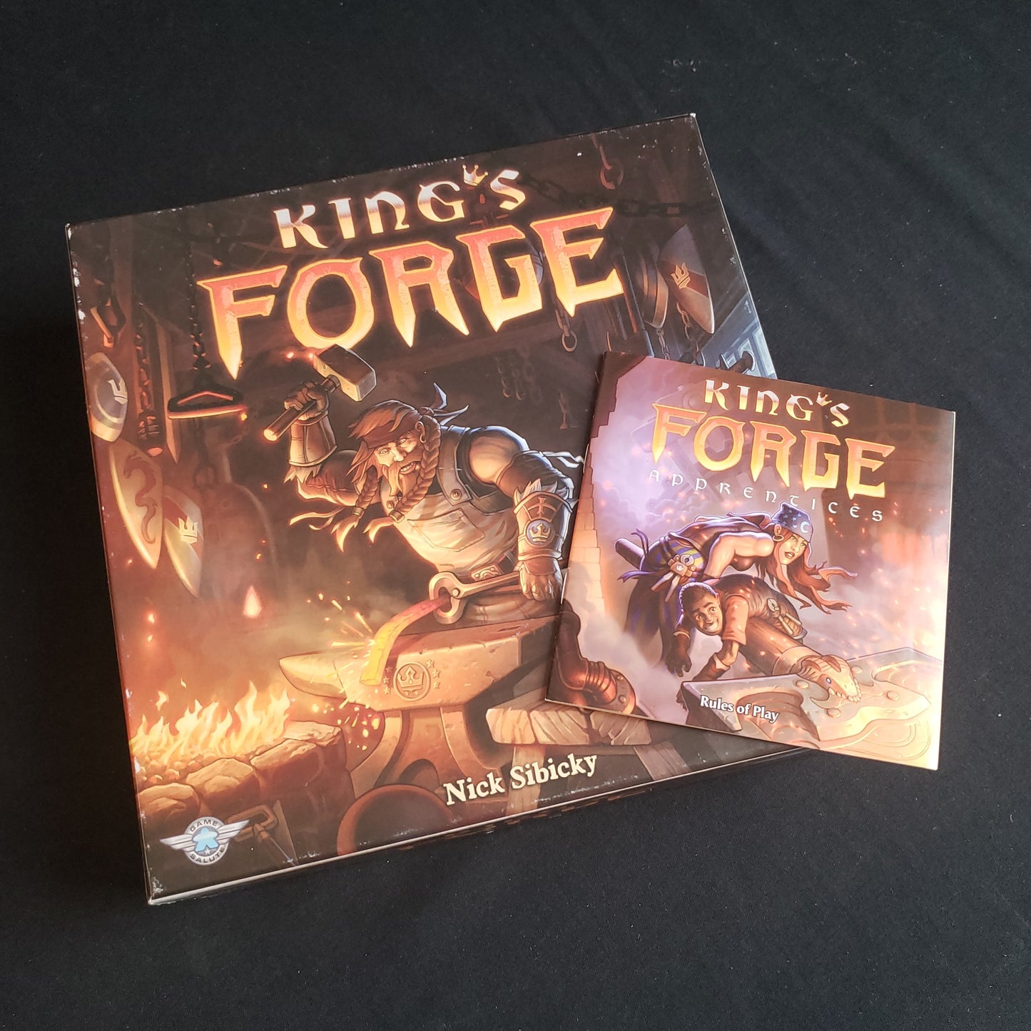 Image shows the front cover of the box of the King's Forge board game, with the instructions for the Apprentices expansion sitting on top of it