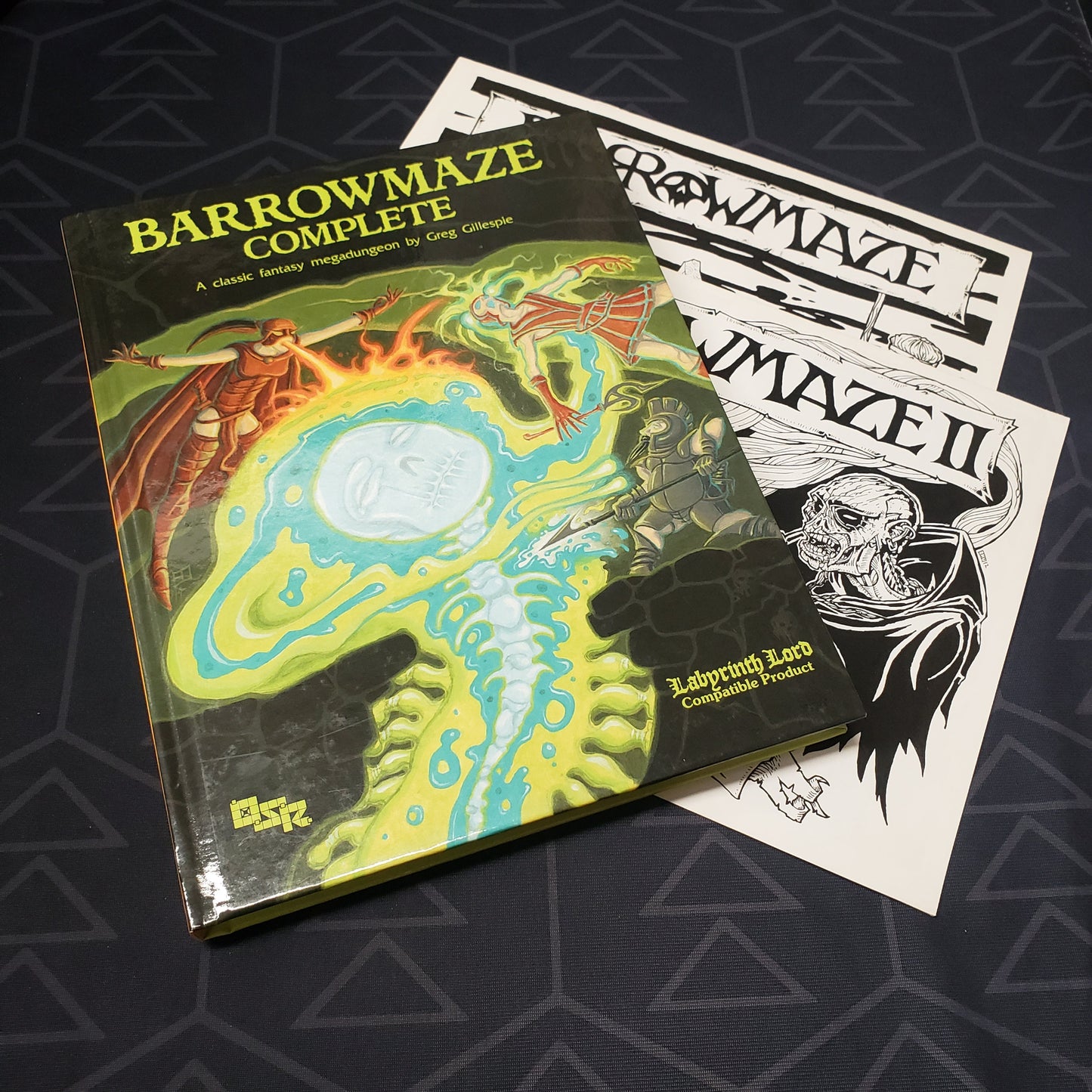 Image shows the front cover of the Barrowmaze Complete book for the roleplaying game Labyrinth Lord, along with two illustration book fanned out