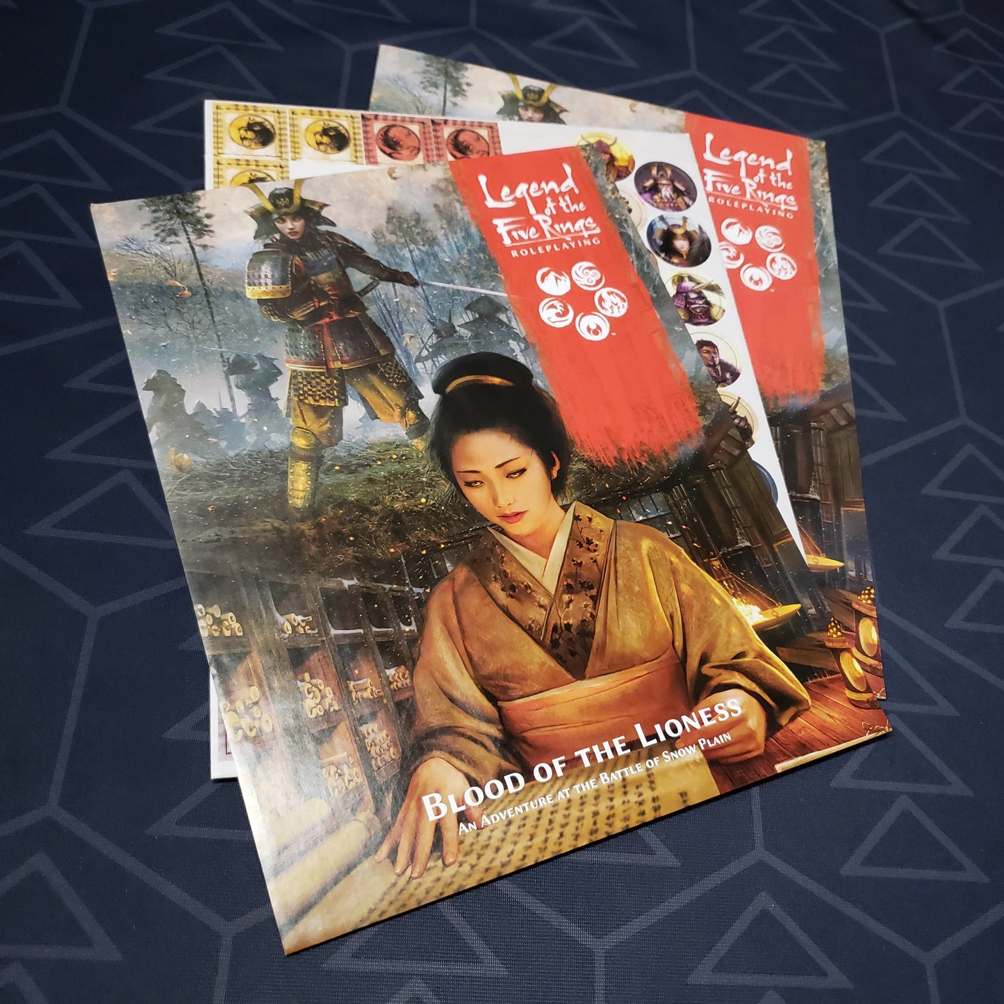 Image shows the fanned-out front cover, token sheet and module of the Blood of the Lioness book for the roleplaying game Legend of the Five Rings: Fifth Edition