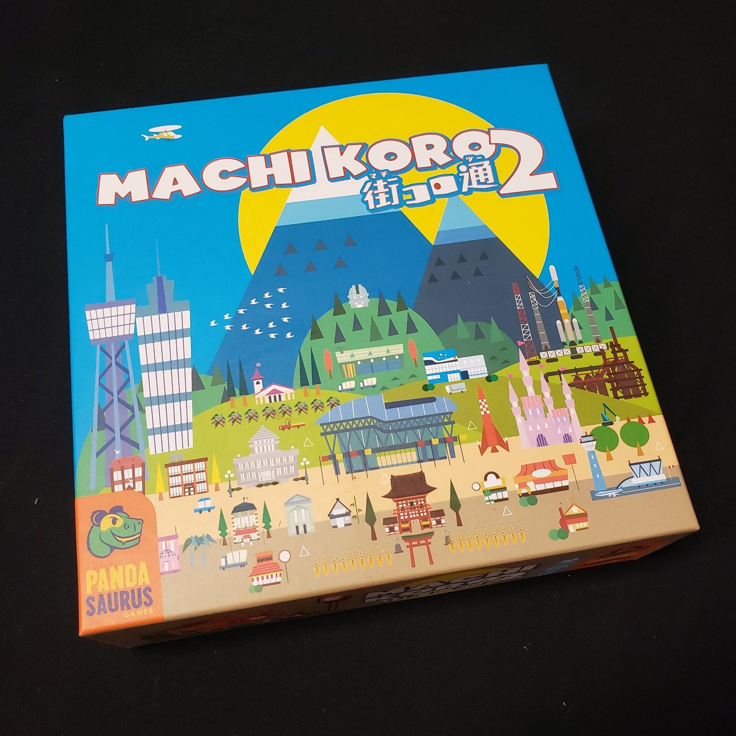 Image shows the front cover of the box of the Machi Koro 2 board game