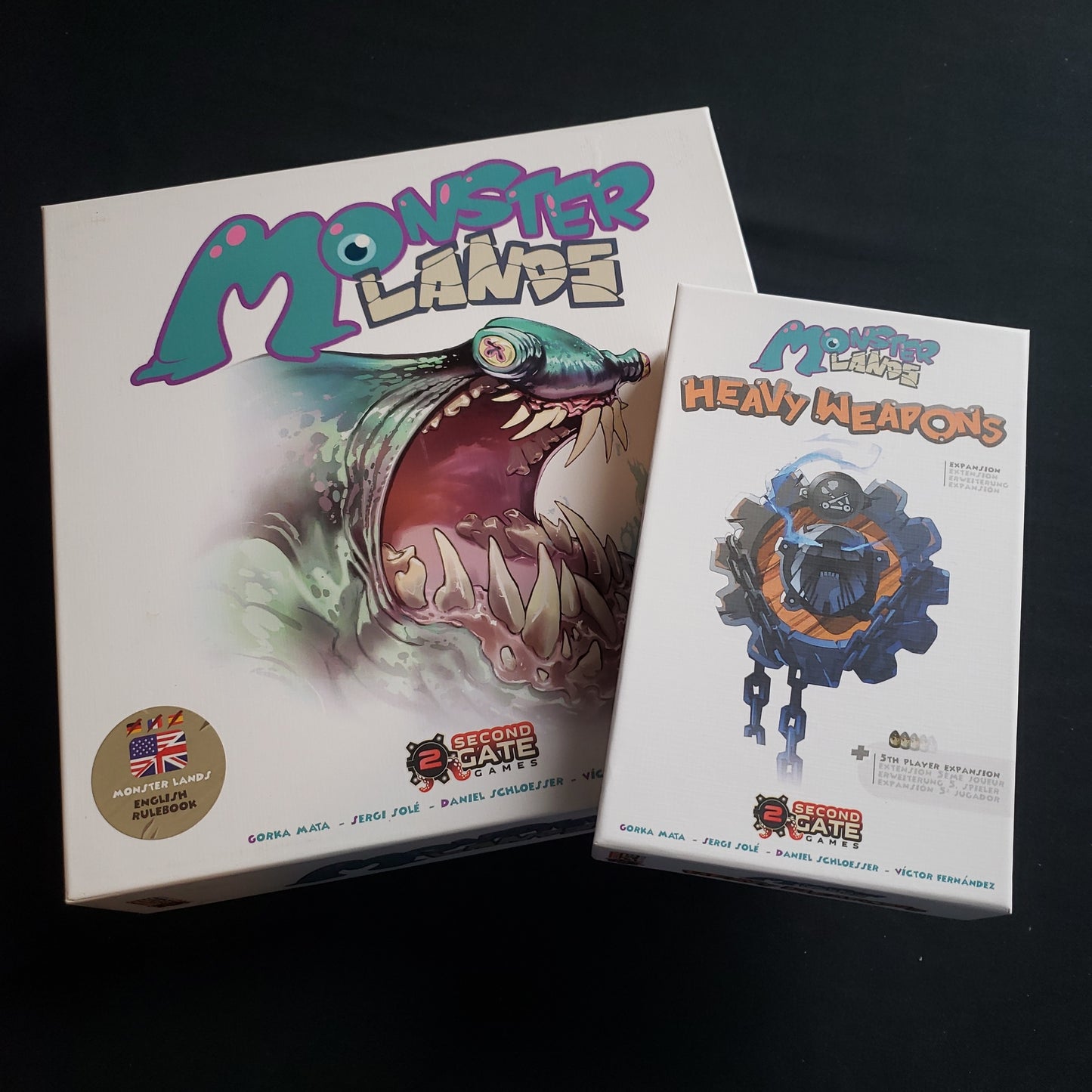 Image shows the front cover of the box of the Monster Lands board game, along with the box for the Heavy Weapons expansion sitting on top of it