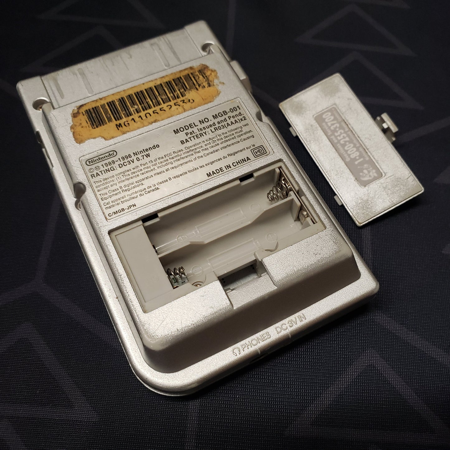 Image shows the back of a Nintendo Game Boy Pocket gaming console with the battery cover sitting beside it