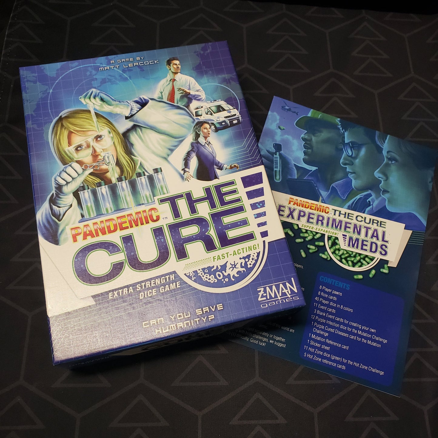 Image shows the front cover of the box of the Pandemic: The Cure board game sitting on top of the instructions for the Experimental meds expansion