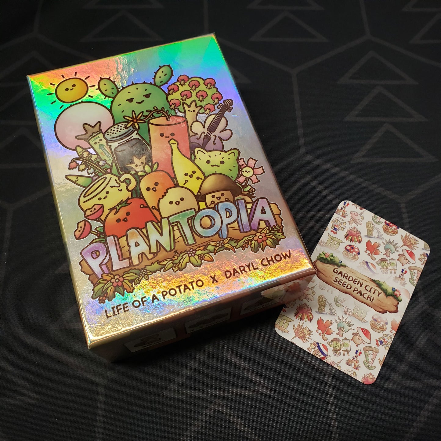 Image shows the front cover of the box for the Plantopia card game sitting next to a card from the Garden City Seed Pack! expansion