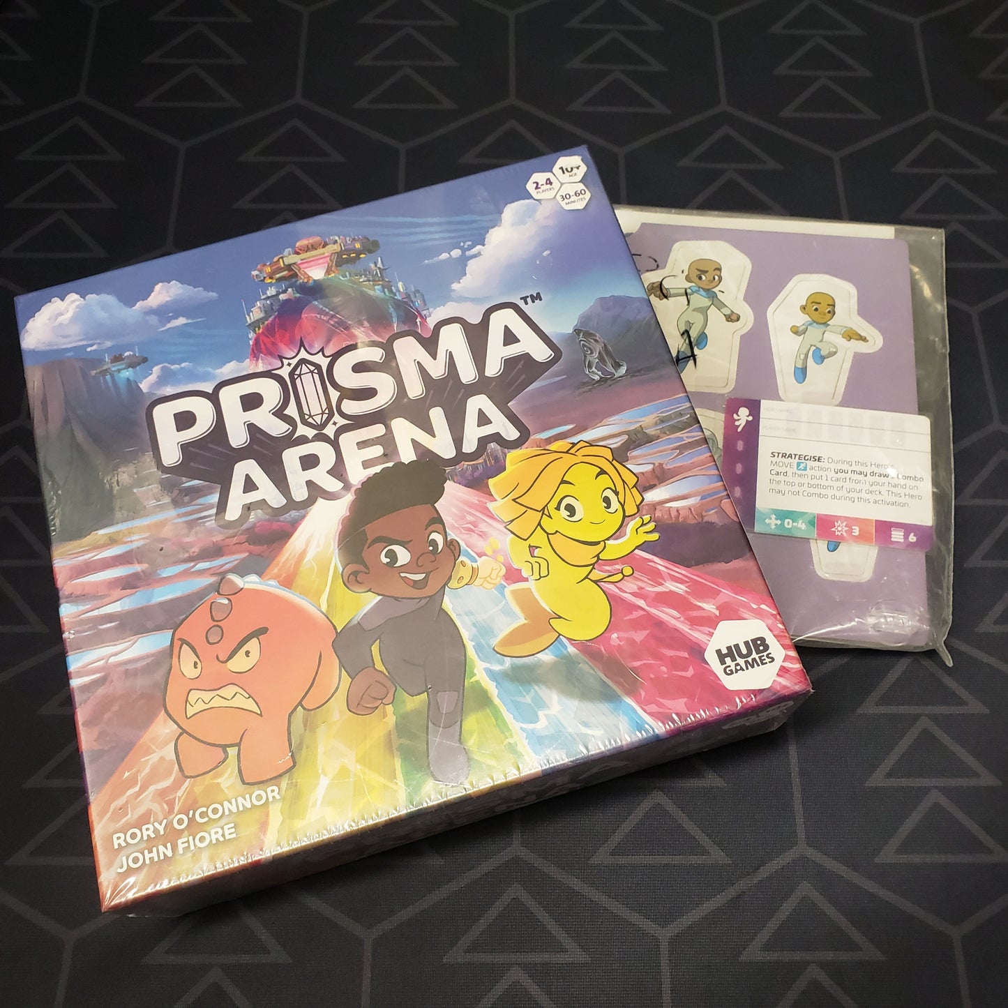 Image shows the front cover of the box of the Prisma Arena board game sitting on top of a punchboard of promo characters