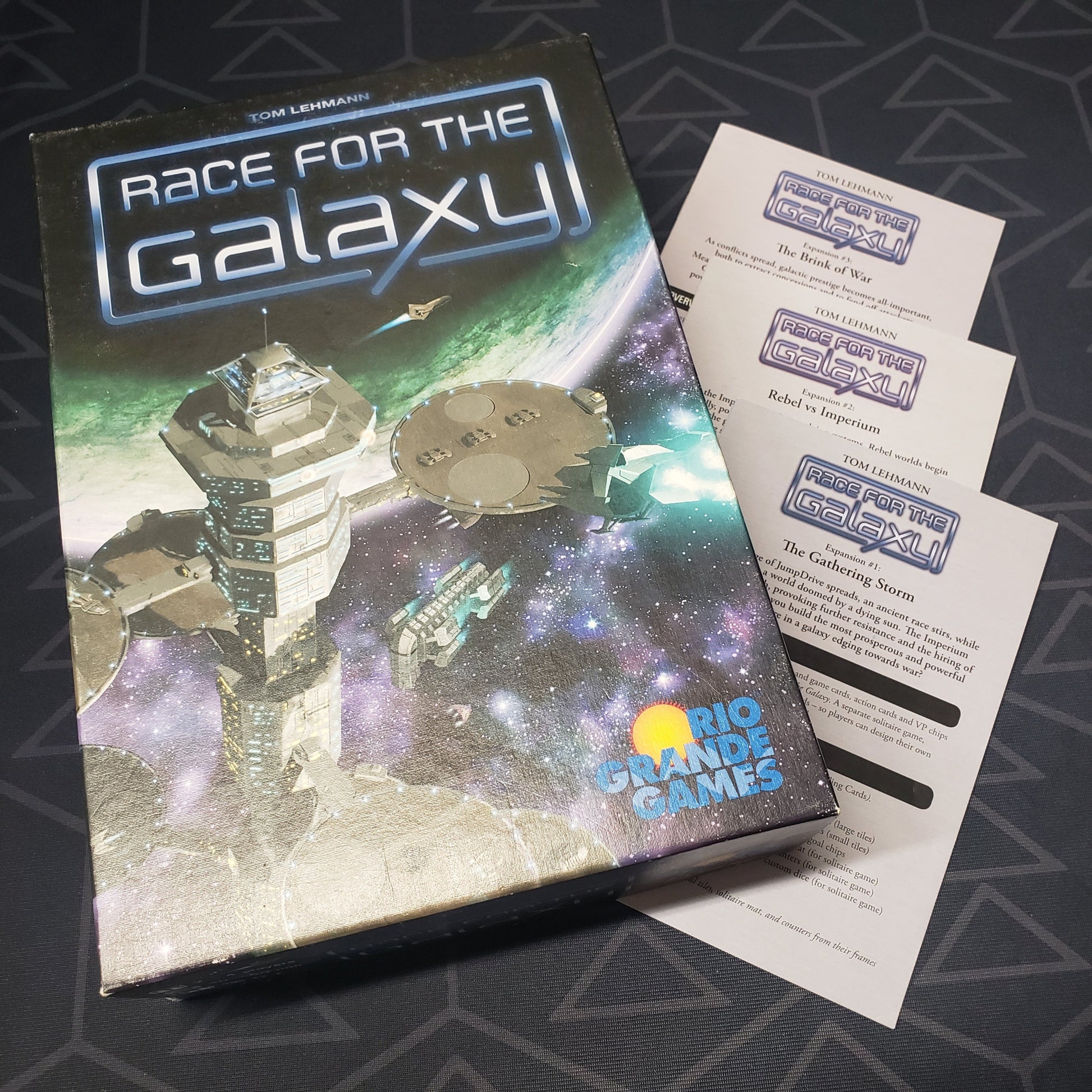 Image shows the front cover of the box for the Race for the Galaxy board game sitting top top of 3 expansion instruction booklets