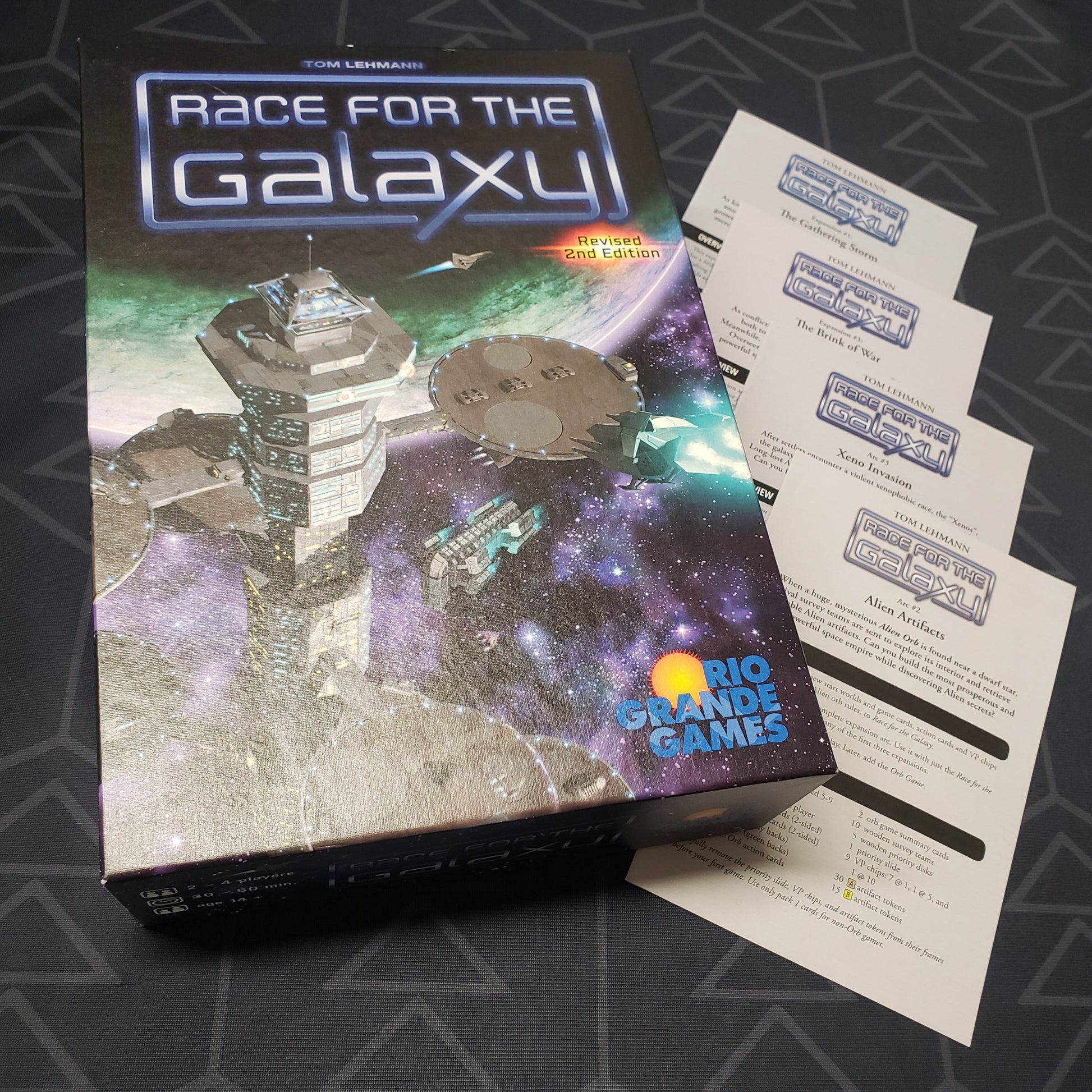 Image shows the front cover of the box for the Race for the Galaxy (Revised Second Edition) board game, with four additional sets of expansion instructions sitting next to it