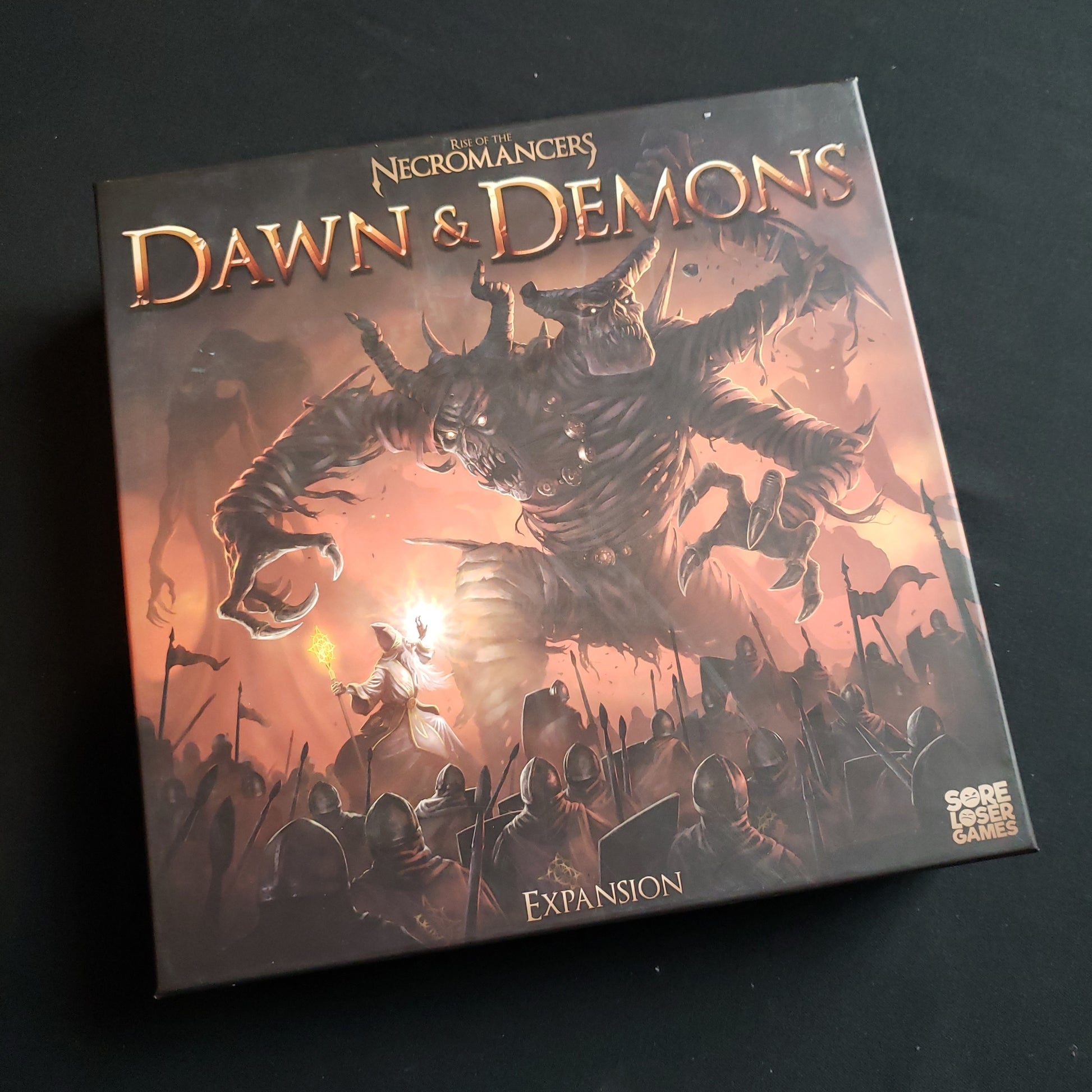 Image shows the front cover of the box of the Dawn & Demons expansion for the board game Rise of the Necromancers