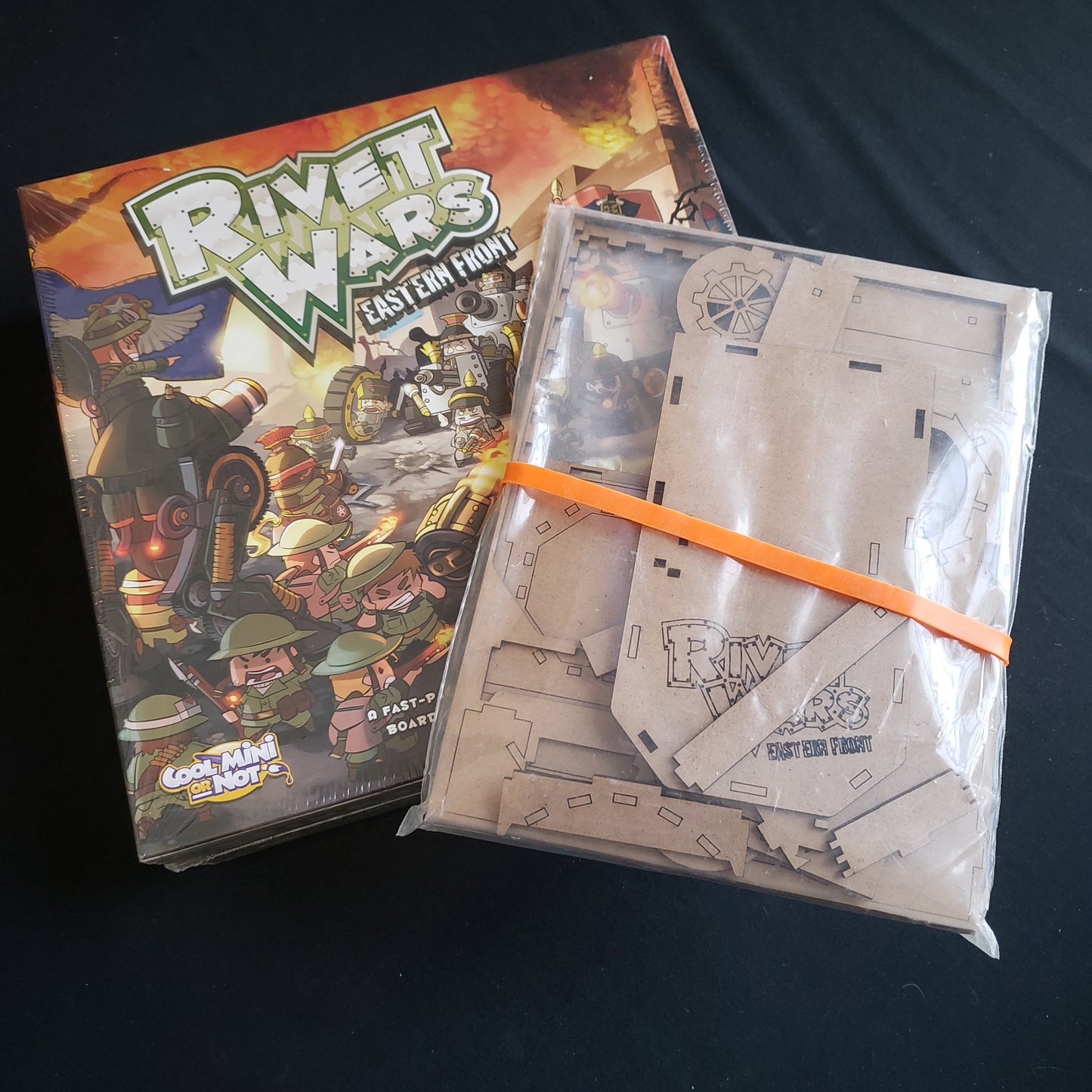 Image shows the front cover of the box of the Rivet Wars board game, with a bag of unassembled wooden storage components sitting on top of it