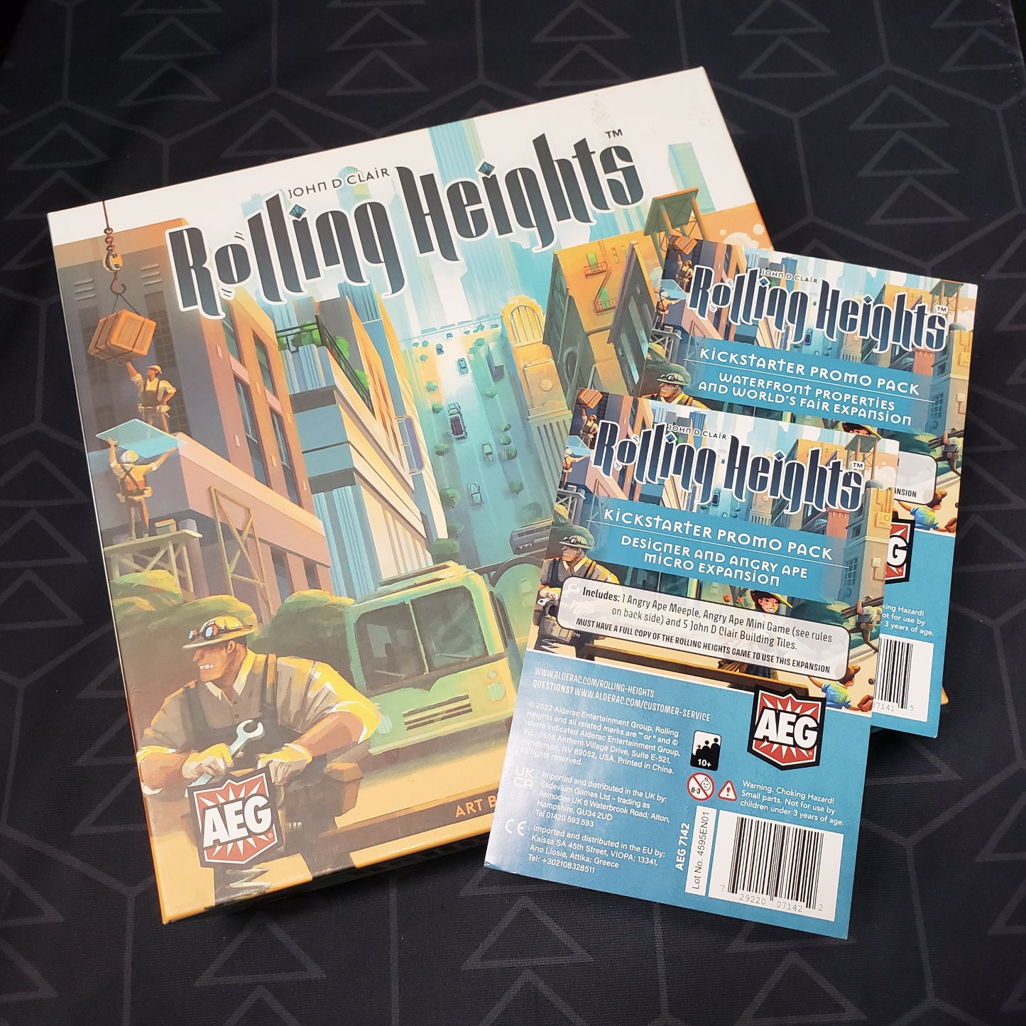 Image shows the front cover of the box of the Rolling Heights board game with the instructions for two Kickstarter promo packs sitting on top of it