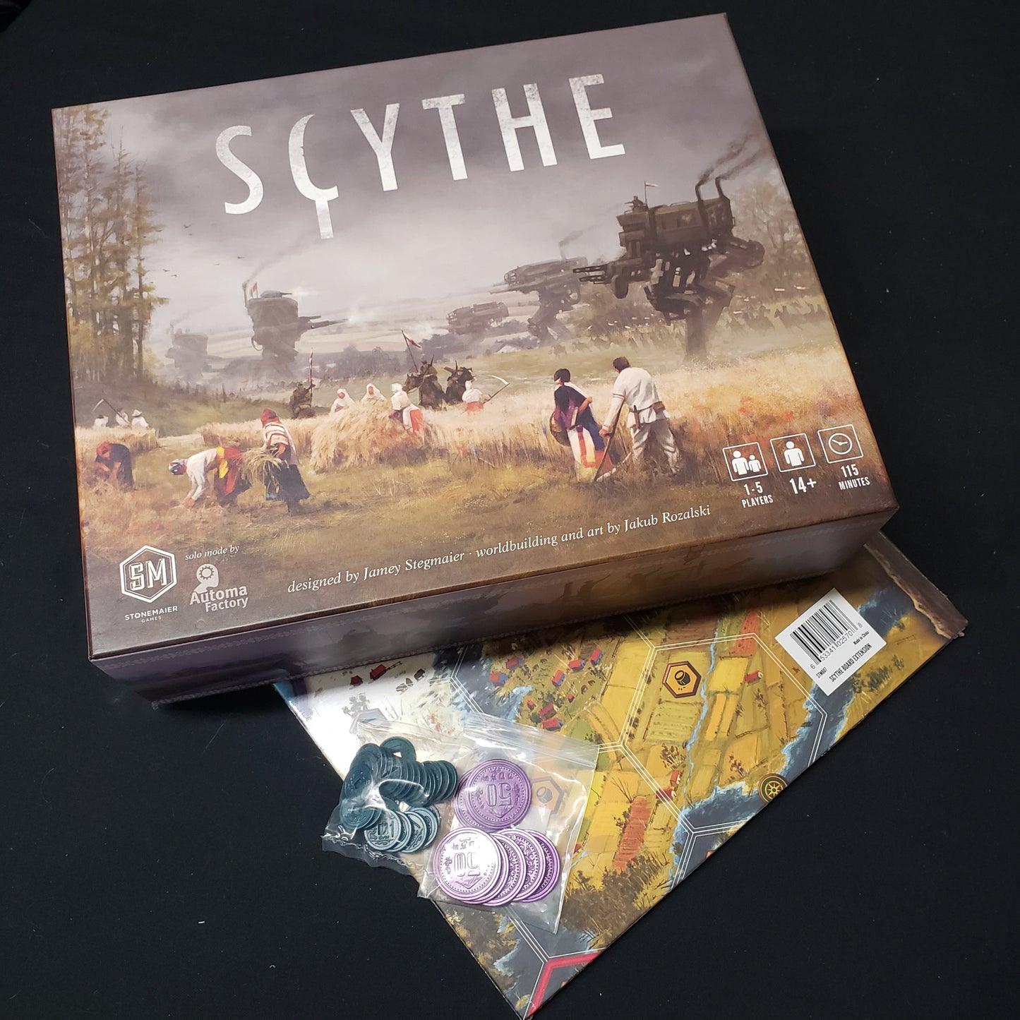 Image shows the front cover of the box of the Scythe board game, sitting on top of the Board Extension and metal coins for the game