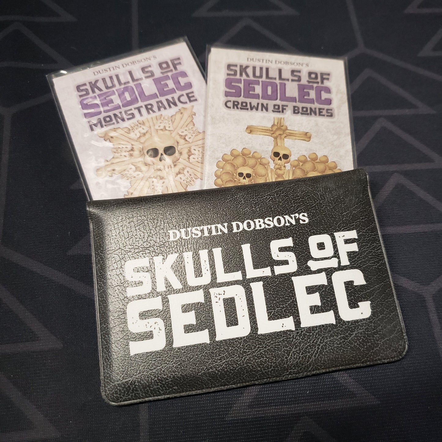 Image shows the front cover of the card wallet of the Skulls of Sedlec card game with the Monstrance and Crown of Bones expansions sitting next to it