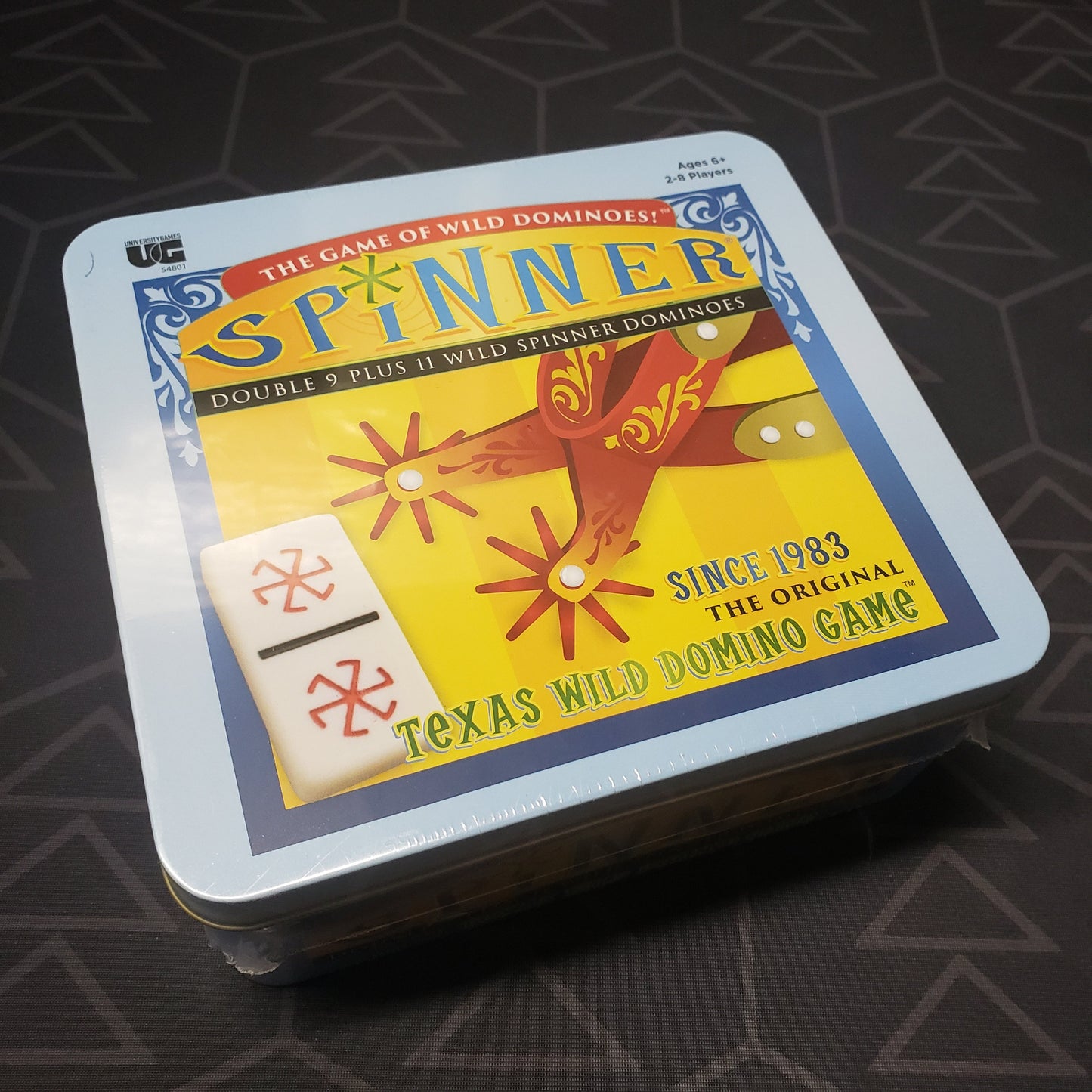 Image shows the front cover of the tin box for the Spinner Dominoes board game