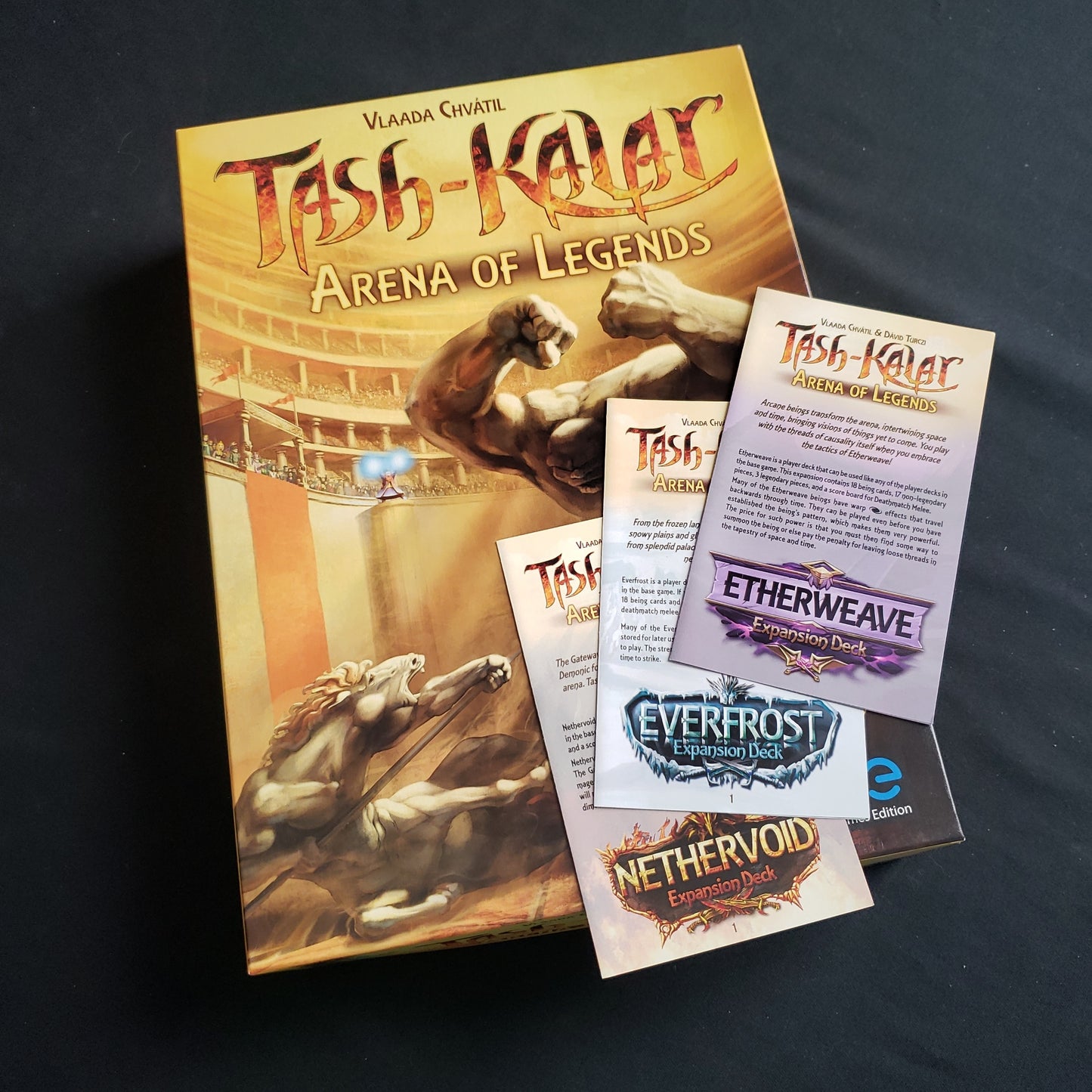 Image shows the front cover of the box of the Tash-Kalar board game, with the instructions for three expansion decks sitting on top of it