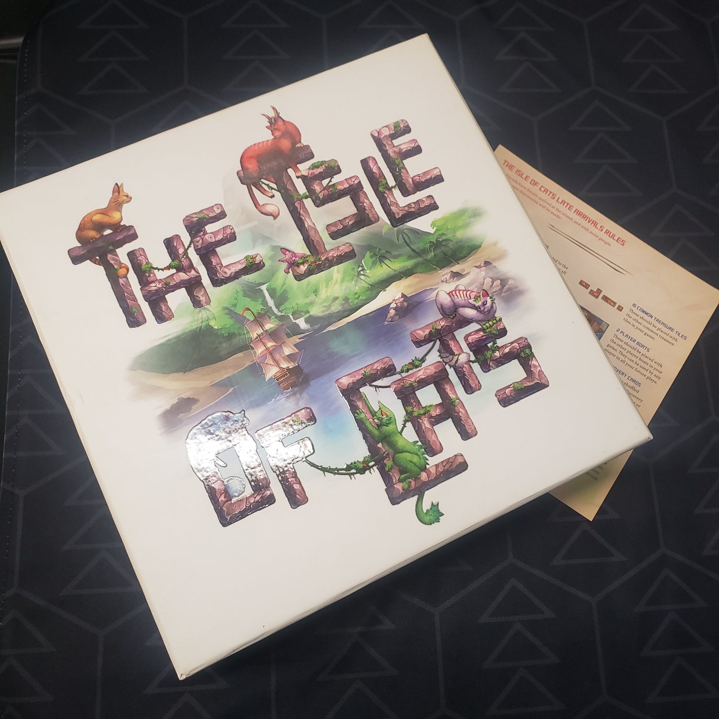 Image shows the front cover of the box of the Isle of Cats board game sitting on top of the instructions for the Late Arrivals expansion