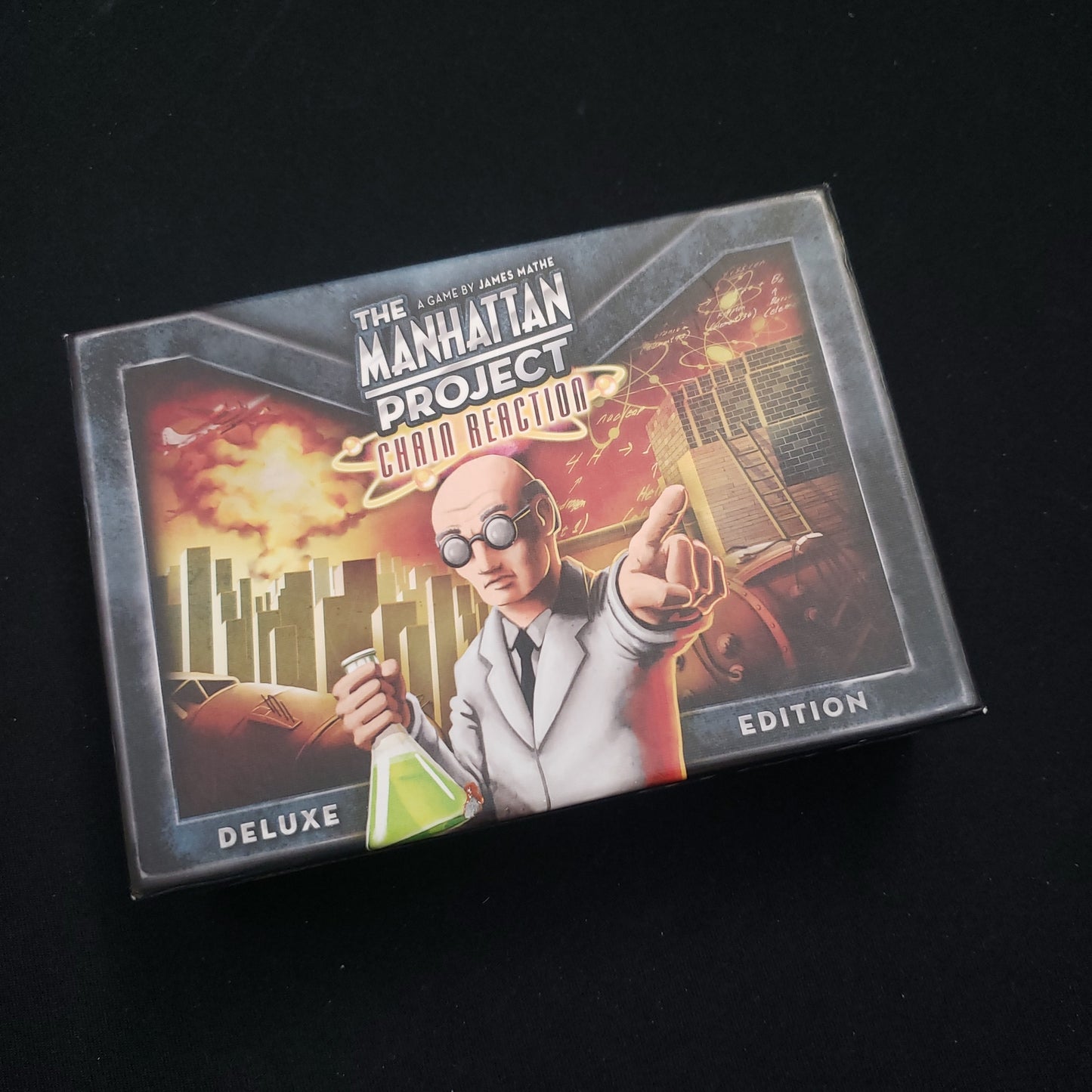 Image shows the front cover of the box of the deluxe edition of the Manhattan Project: Chain Reaction card game