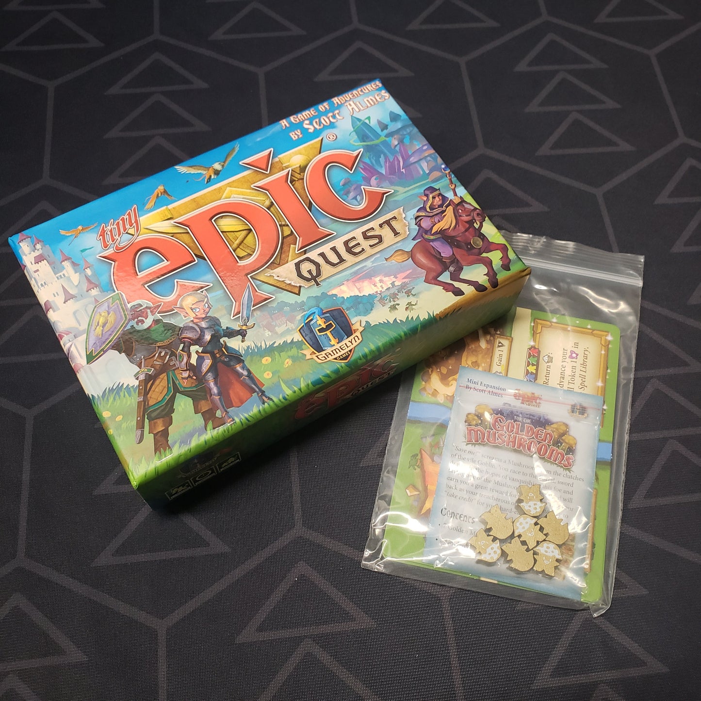 Image shows the front cover of the box for the Tiny Epic Quest board game sitting next to a baggie of expansion components