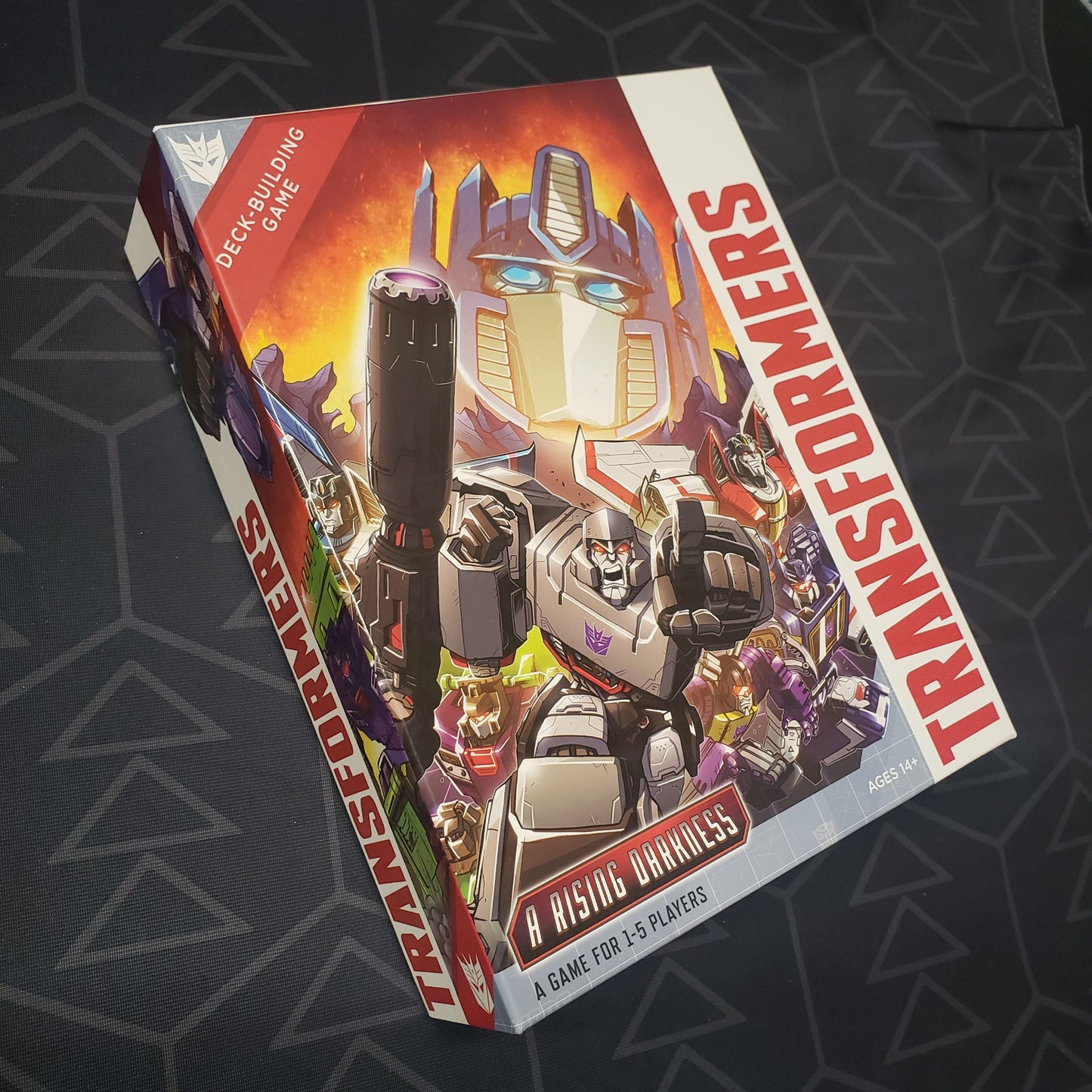 Image shows the front cover of the box of the A Rising Darkness expansion for the card game Transformers Deck-Building Game