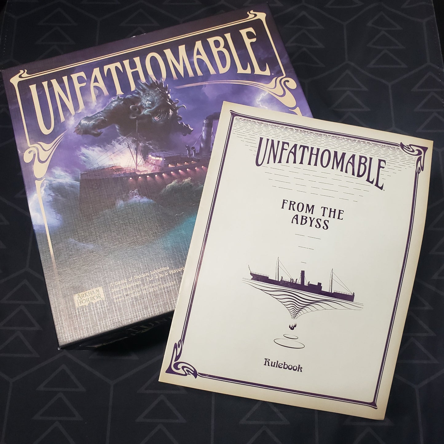 Image shows the front cover of the box of the Unfathomable board game with the instructions for the From The Abyss expansion sitting on top of it
